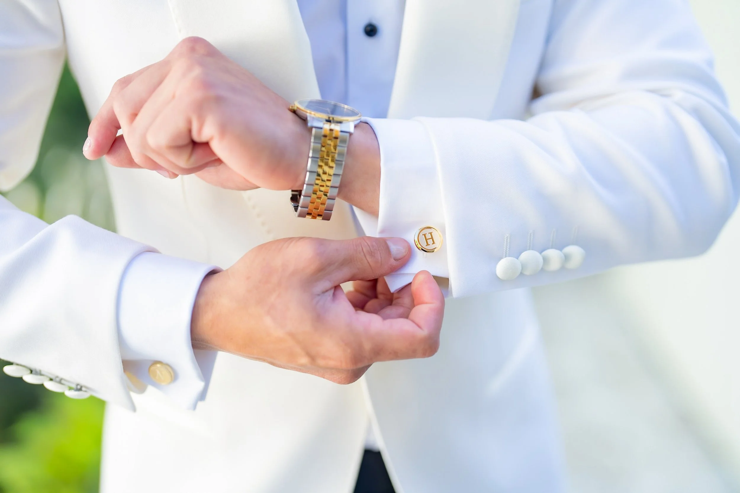 A person in a white suit adjusting their cufflinks and watch, with a gold and silver bracelet, outdoors in a natural setting.
