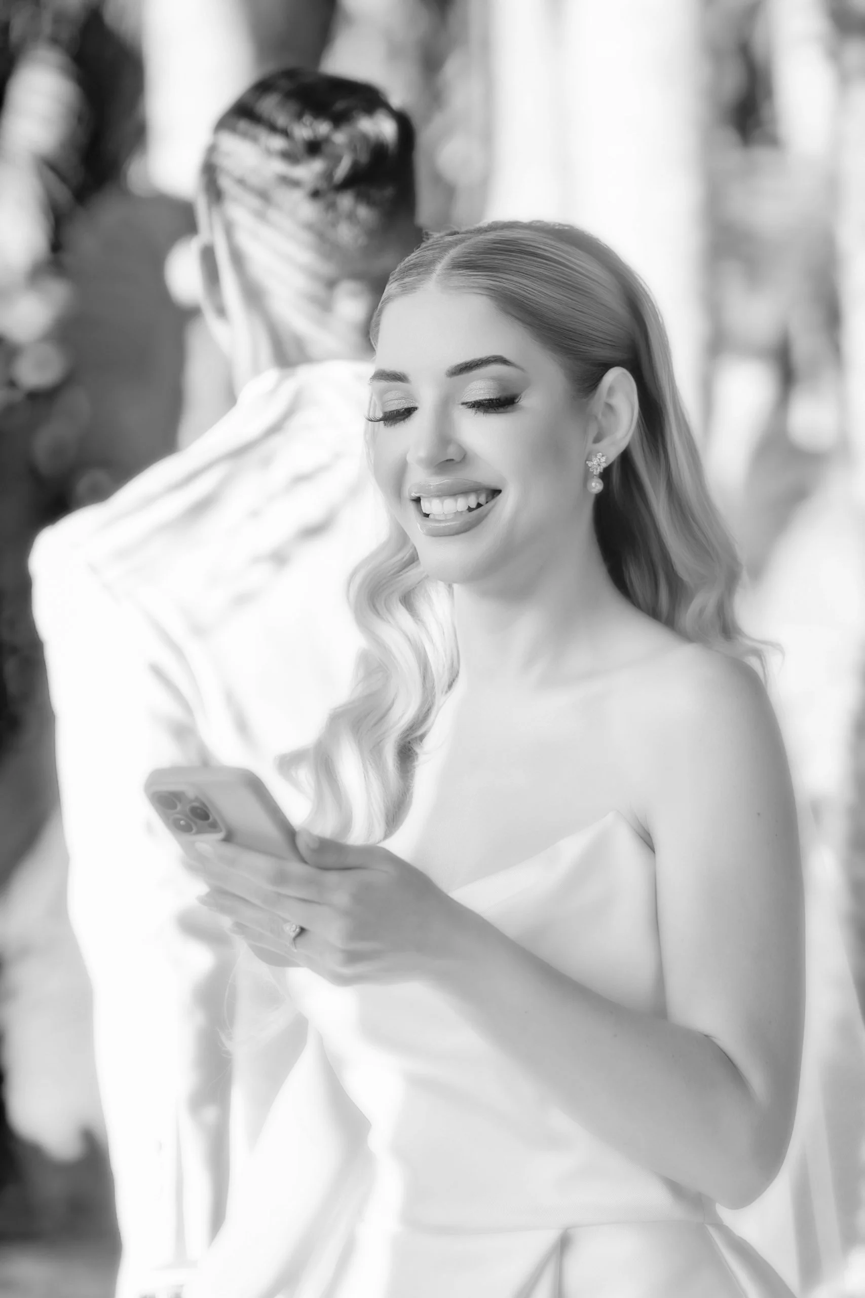 A smiling woman with long wavy hair, wearing earrings and a strapless dress, looking at her phone. Behind her, a man and another woman are blurred in the background, outdoors.