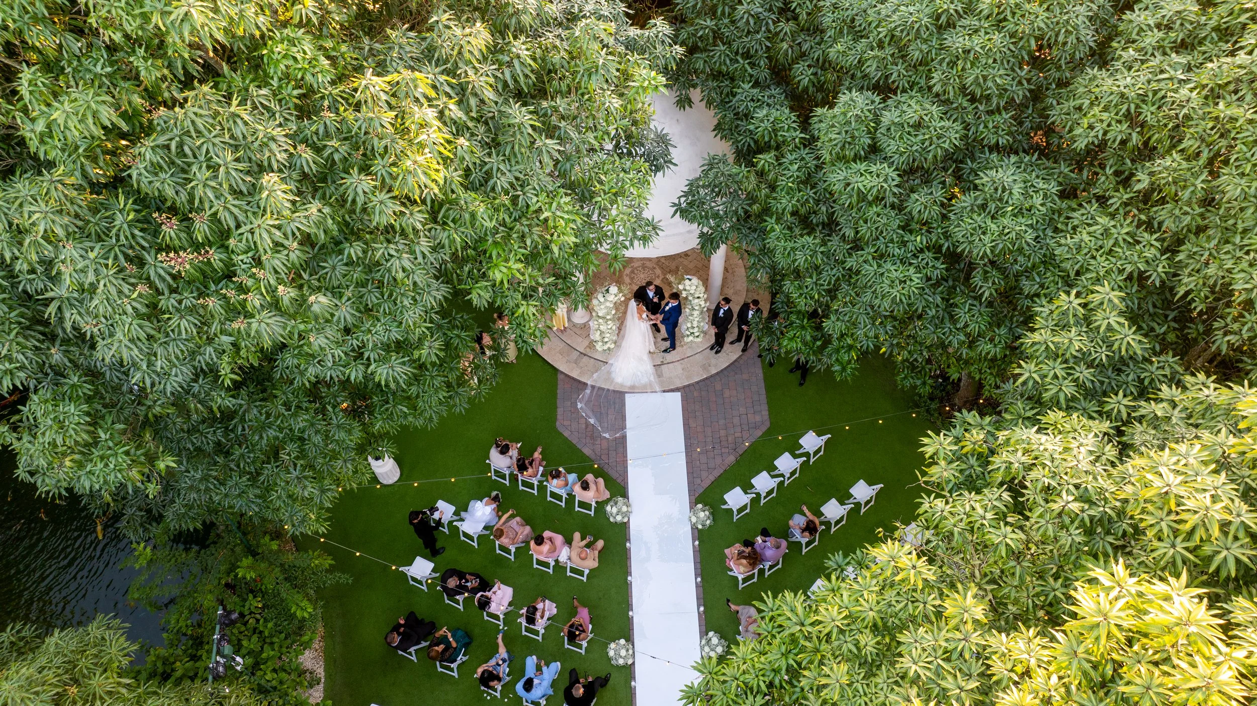 Bird's-eye view of an outdoor wedding ceremony surrounded by lush green trees. The bride and groom stand under an arch decorated with white flowers, with guests seated on white chairs on a grassy area. Some guests are standing and socializing nearby.