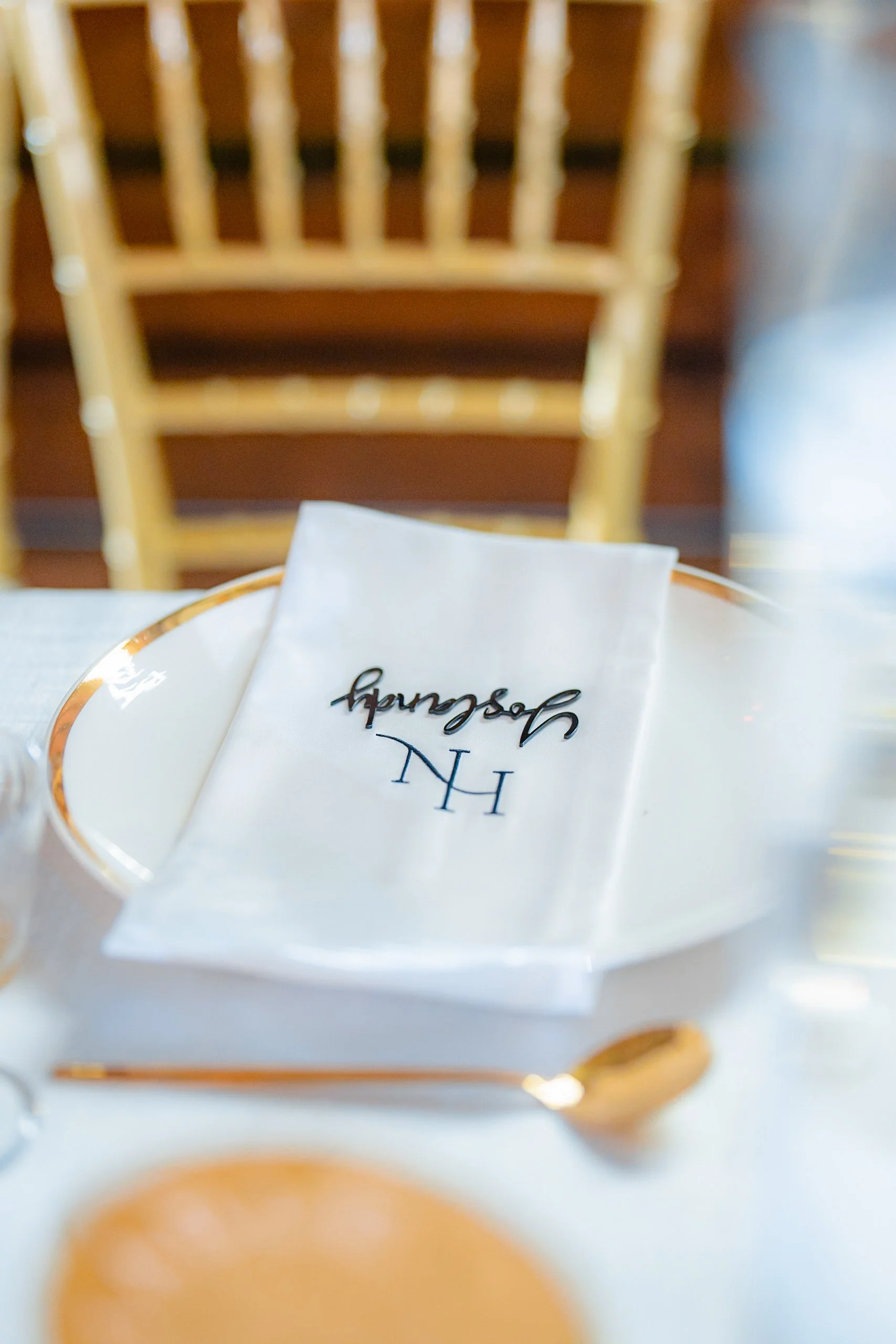 A table setting with a white napkin featuring the embroidered initials "H" and "H" and the word "Hampshire" in black script. There is a gold-rimmed plate, a gold spoon, and a partial view of a glass and a decorative orange slice on the table. A woode