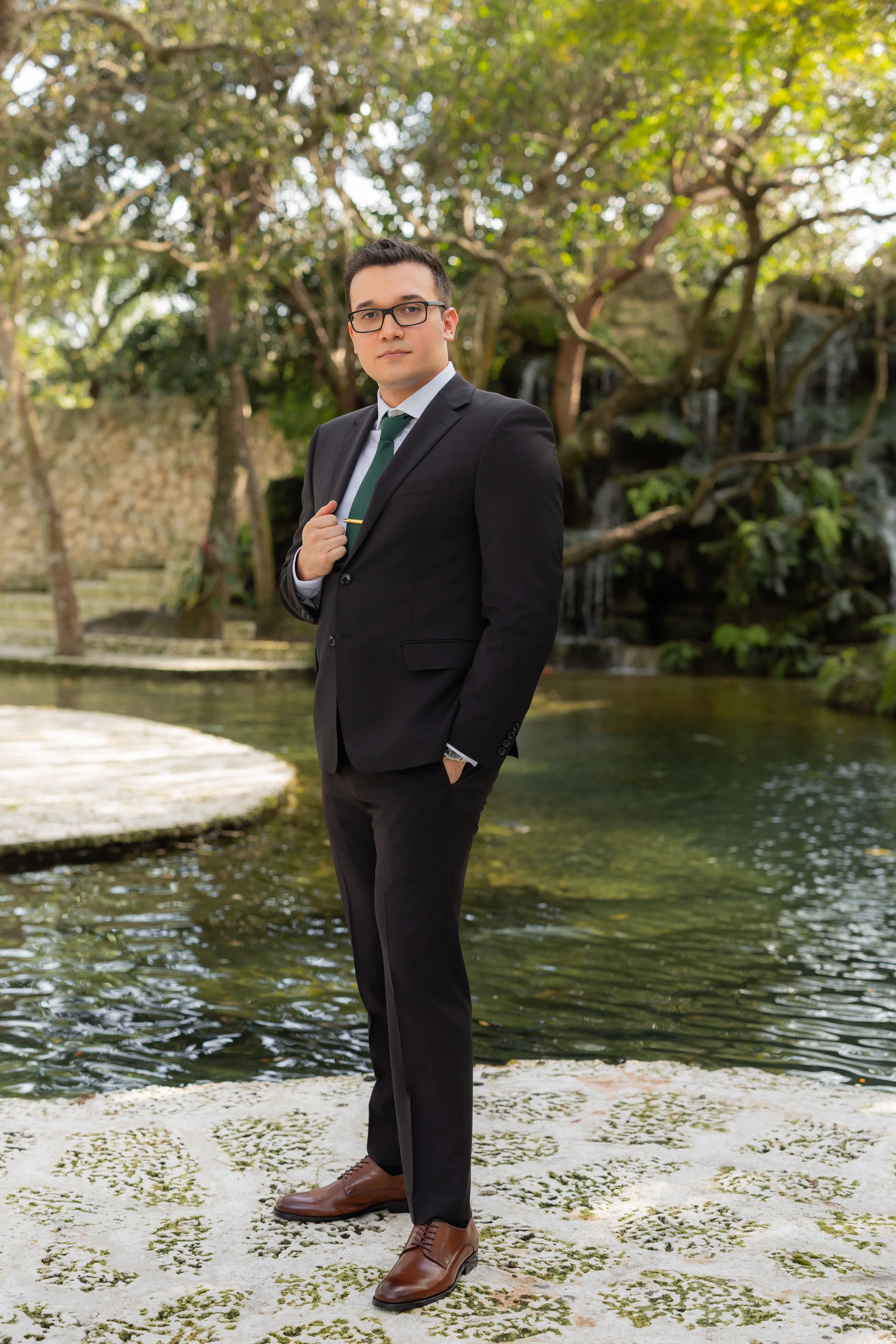 A young man in a black suit, white shirt, green tie, and brown dress shoes posing outdoors by a pond with rocks, trees, and a waterfall in the background.