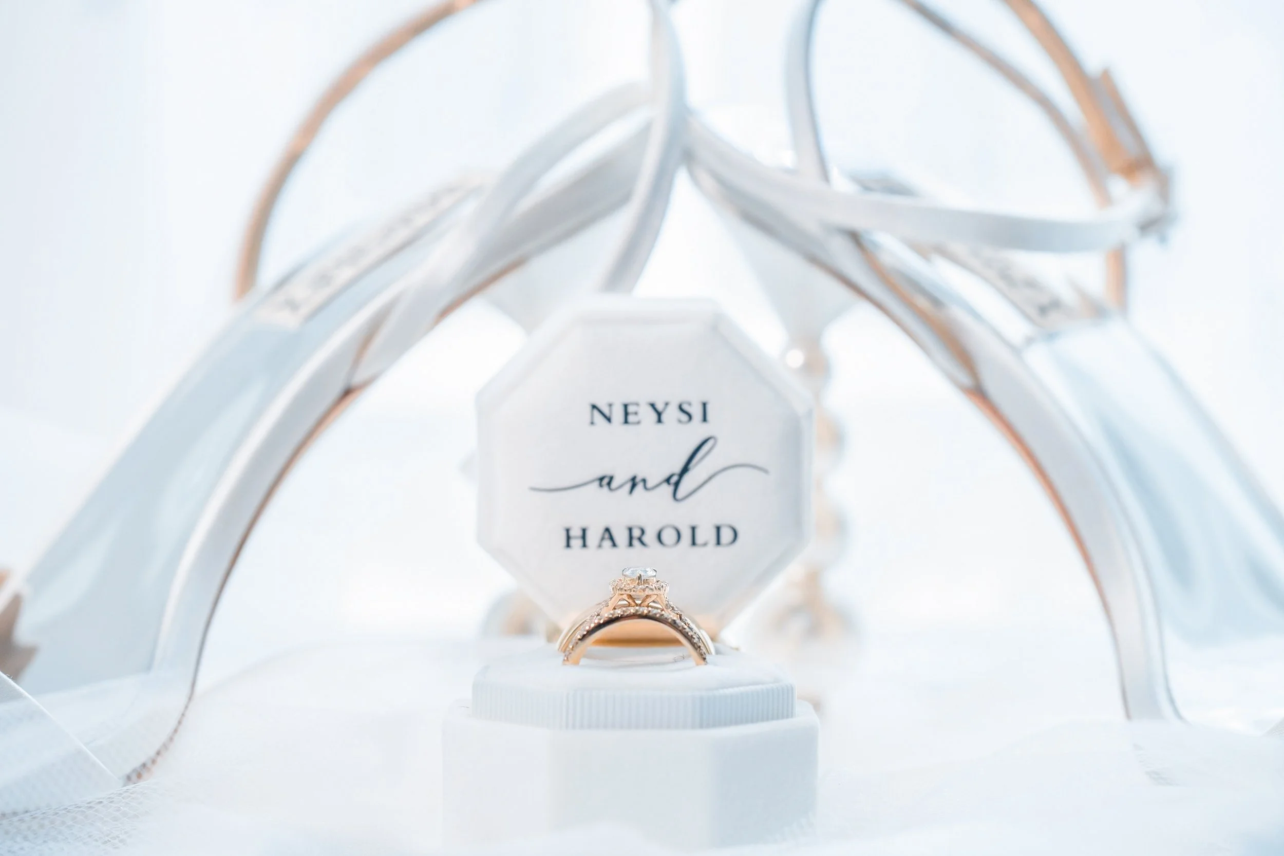 Wedding rings in front of a white arch with a sign that has the names 'Neysi and Harold' written on it.