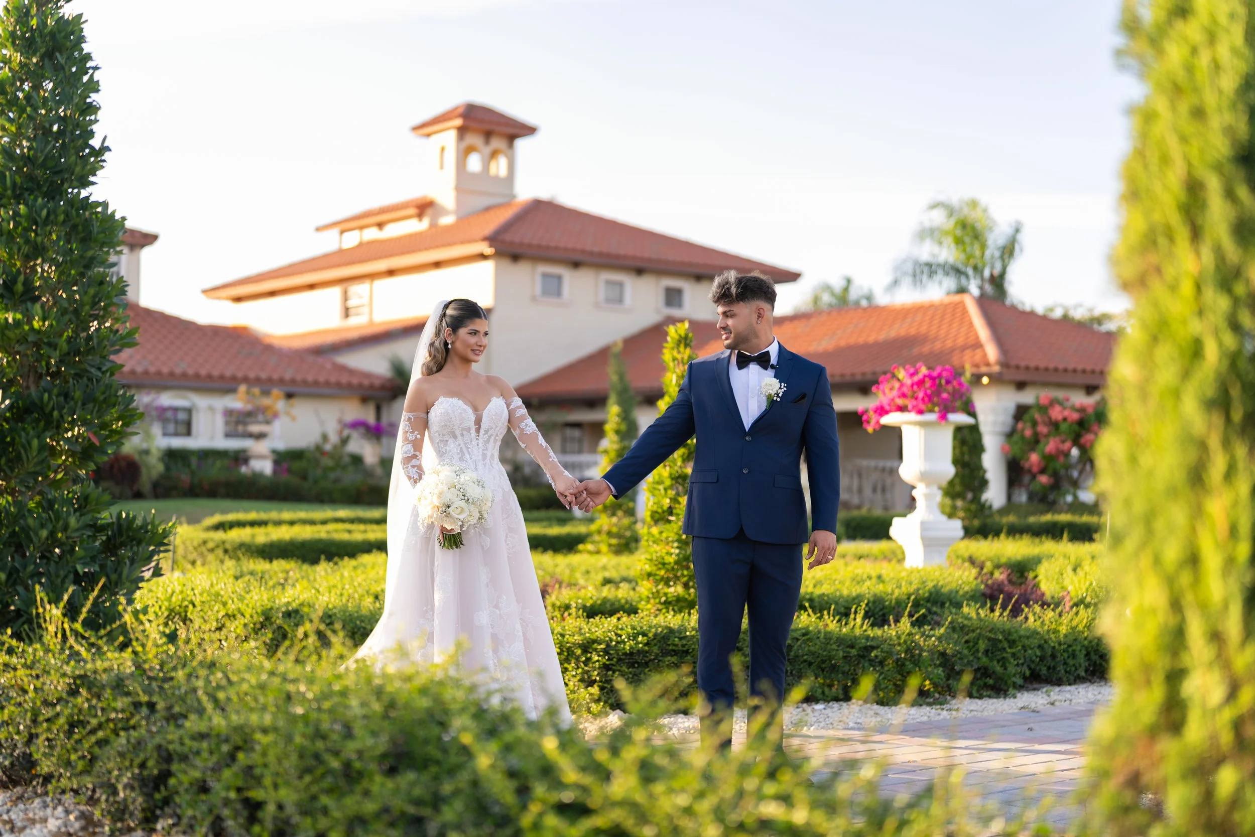 A bride and groom holding hands in a garden with a large house in the background.