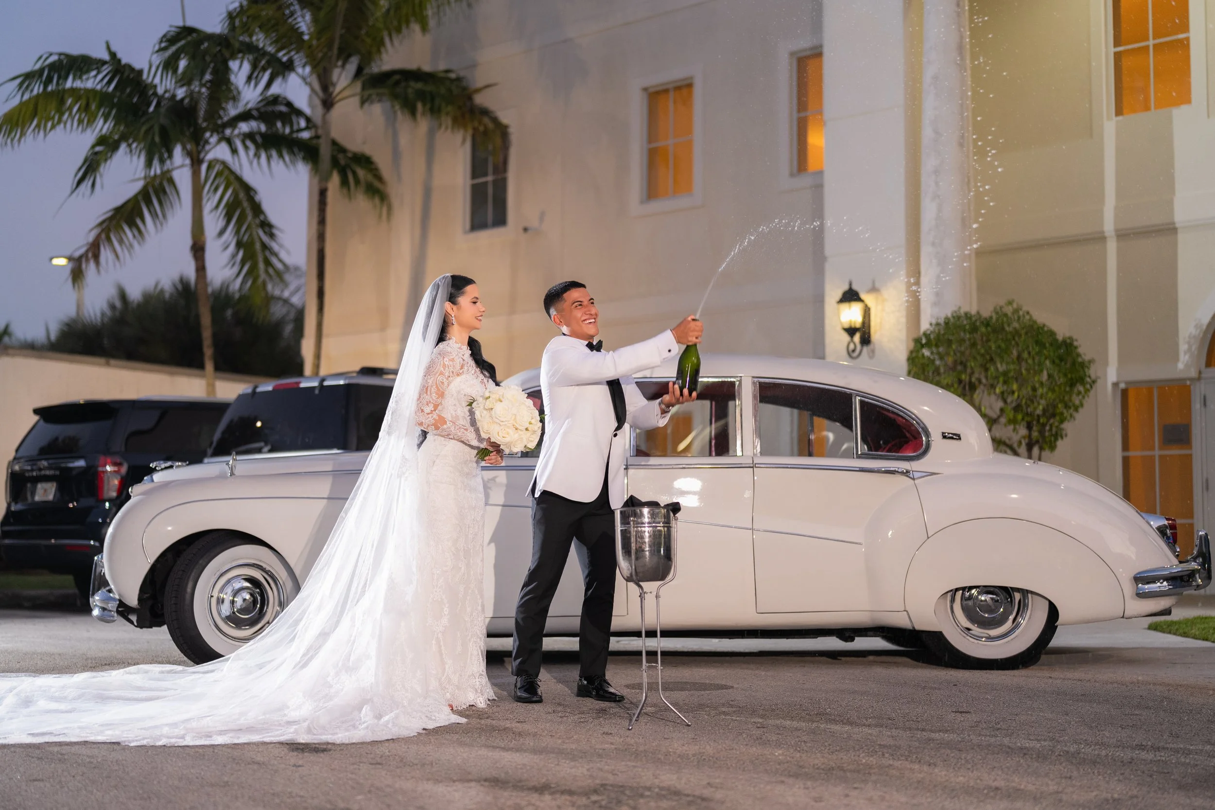 Bride and groom celebrating outside near a vintage white car, with the groom opening a bottle of champagne as the bride looks on smiling, during their wedding night.