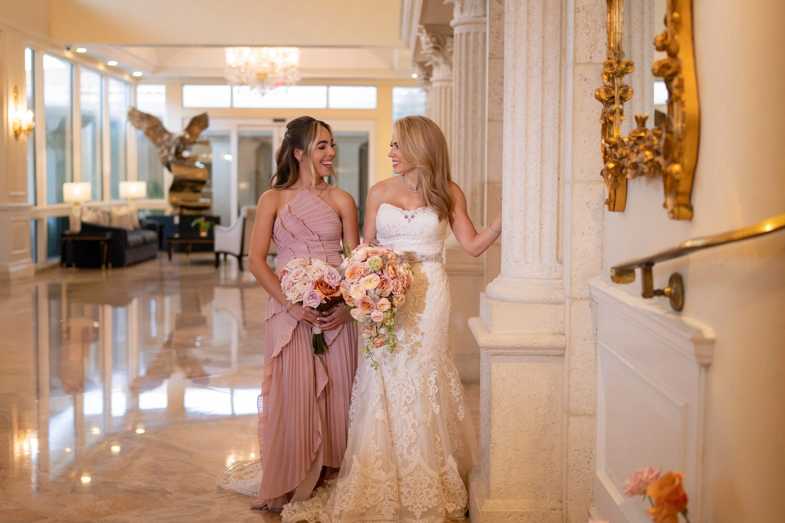 Two women, one in a white wedding dress and the other in a pink dress, smiling and holding bouquets, in an elegant, well-lit hotel lobby or reception area with marble floors and ornate decor.