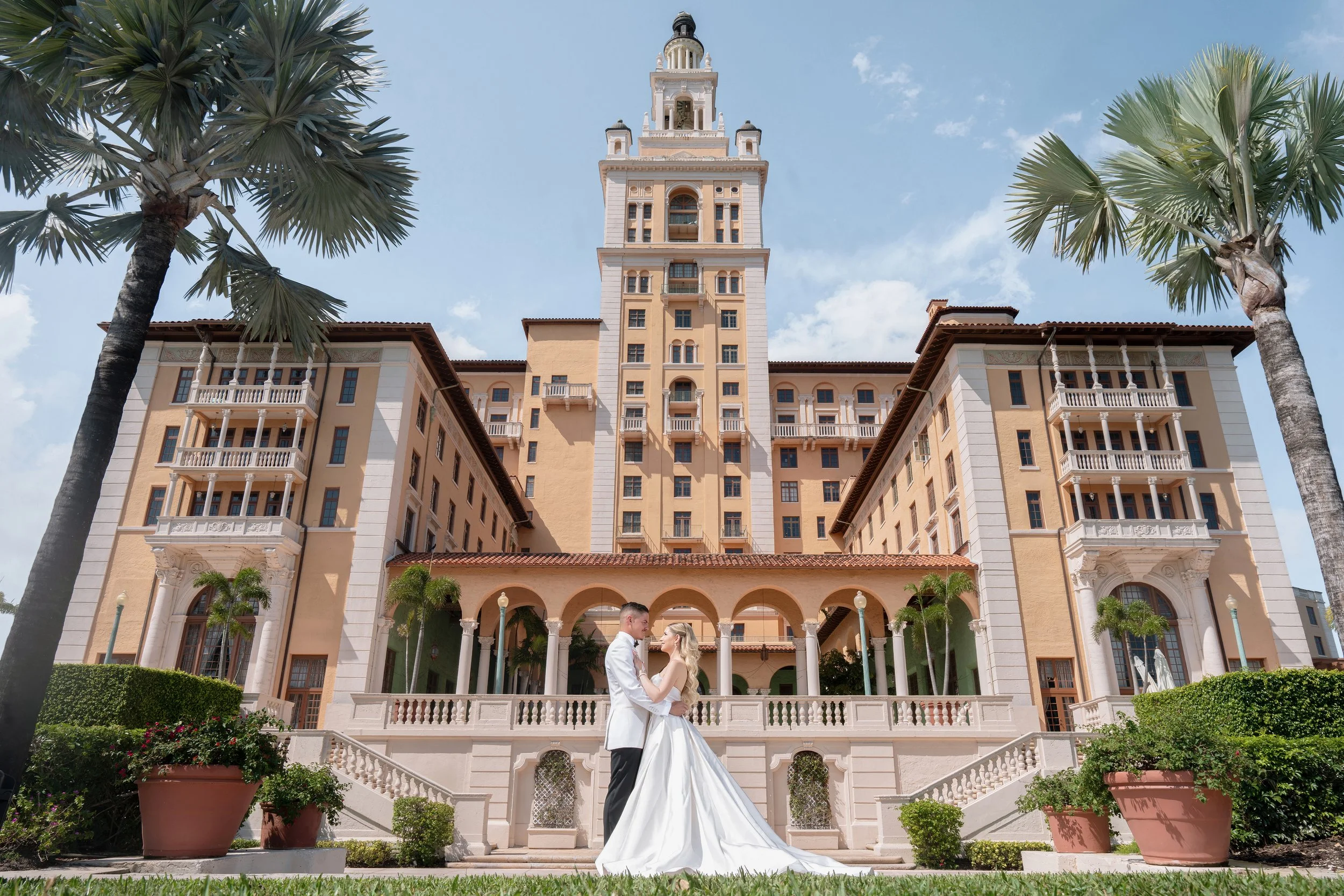 A bride and groom in wedding attire stand embracing in front of a large, elegant, castle-like building with towers and balconies, surrounded by palm trees and potted plants under a partly cloudy sky.