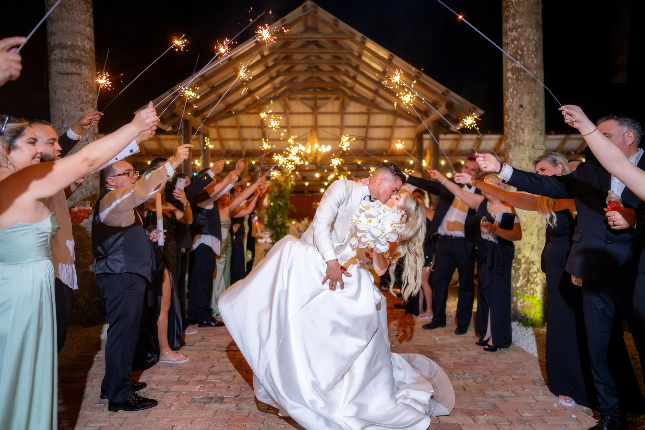 Bride and groom kiss under sparklers during wedding celebration at night, surrounded by guests holding sparklers.