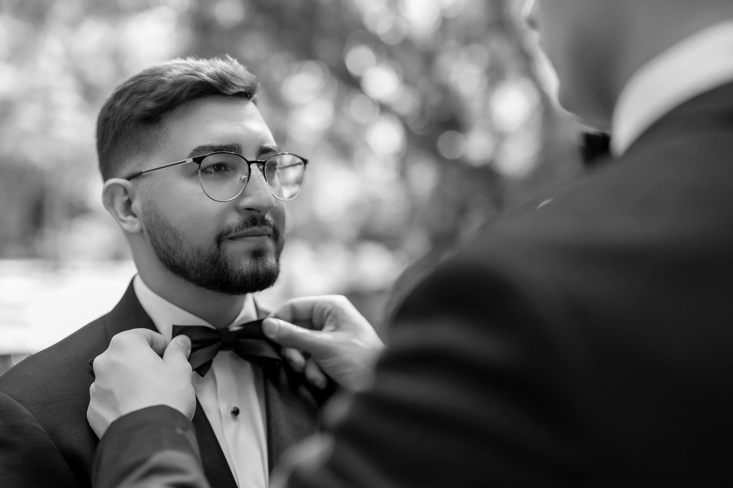 A man with glasses, a beard, and short hair wearing a tuxedo and bow tie, getting assistance from another person adjusting his bow tie outdoors.