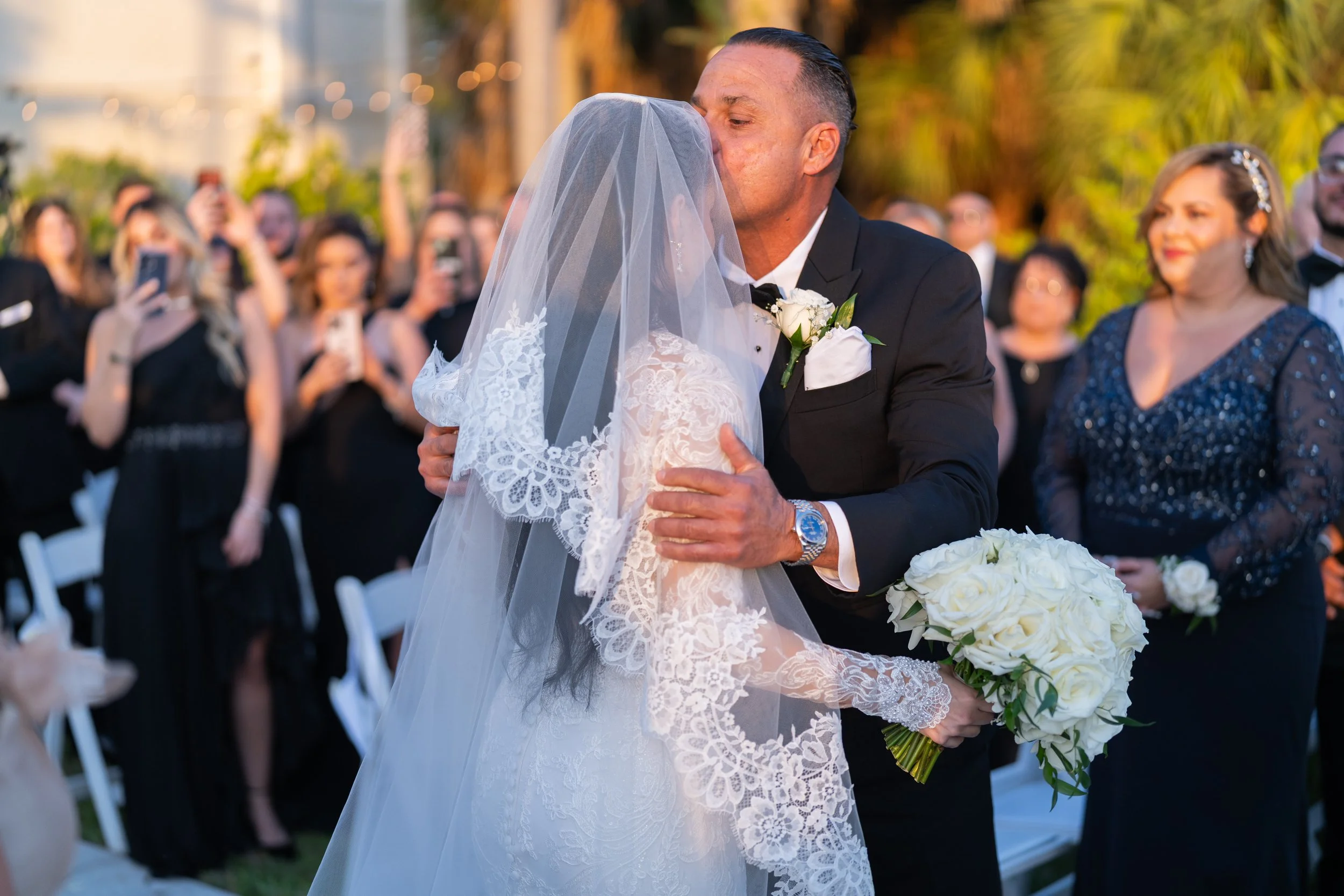 A bride and groom share a kiss during their outdoor wedding ceremony, with guests in the background taking photos and smiling.