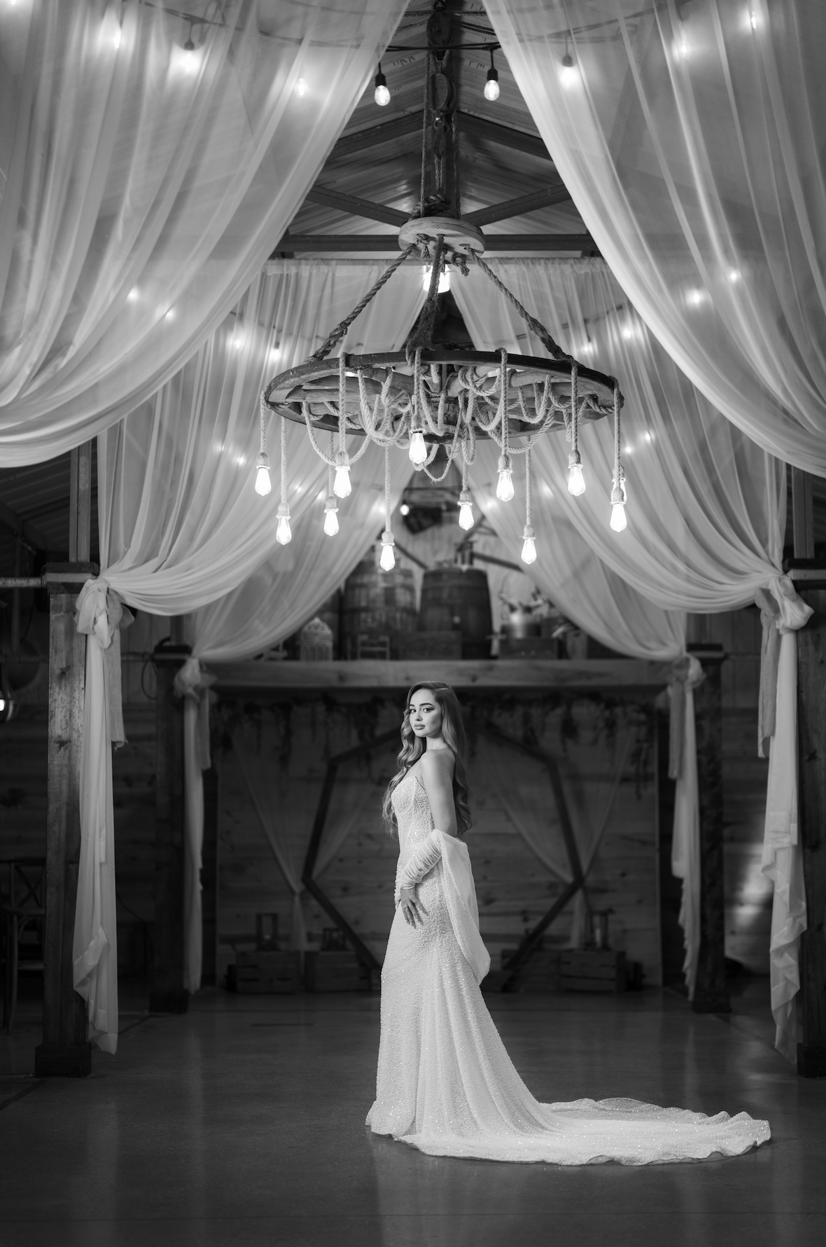 A woman in a long, elegant wedding dress standing in a decorated rustic barn, with draped curtains and a rustic chandelier overhead, in black and white.