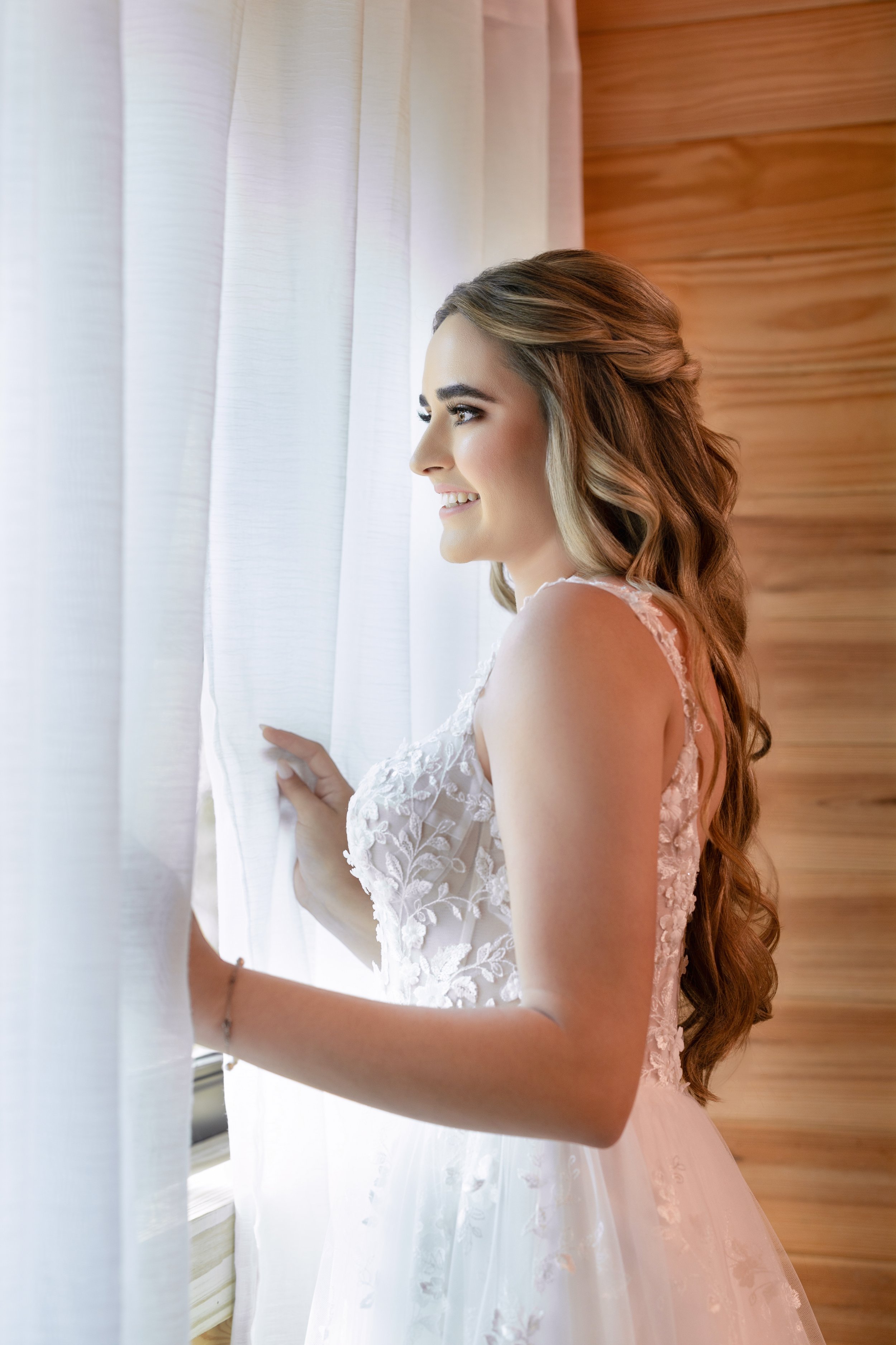 A young woman with long, wavy hair looking out a window, smiling, in a white lace dress.