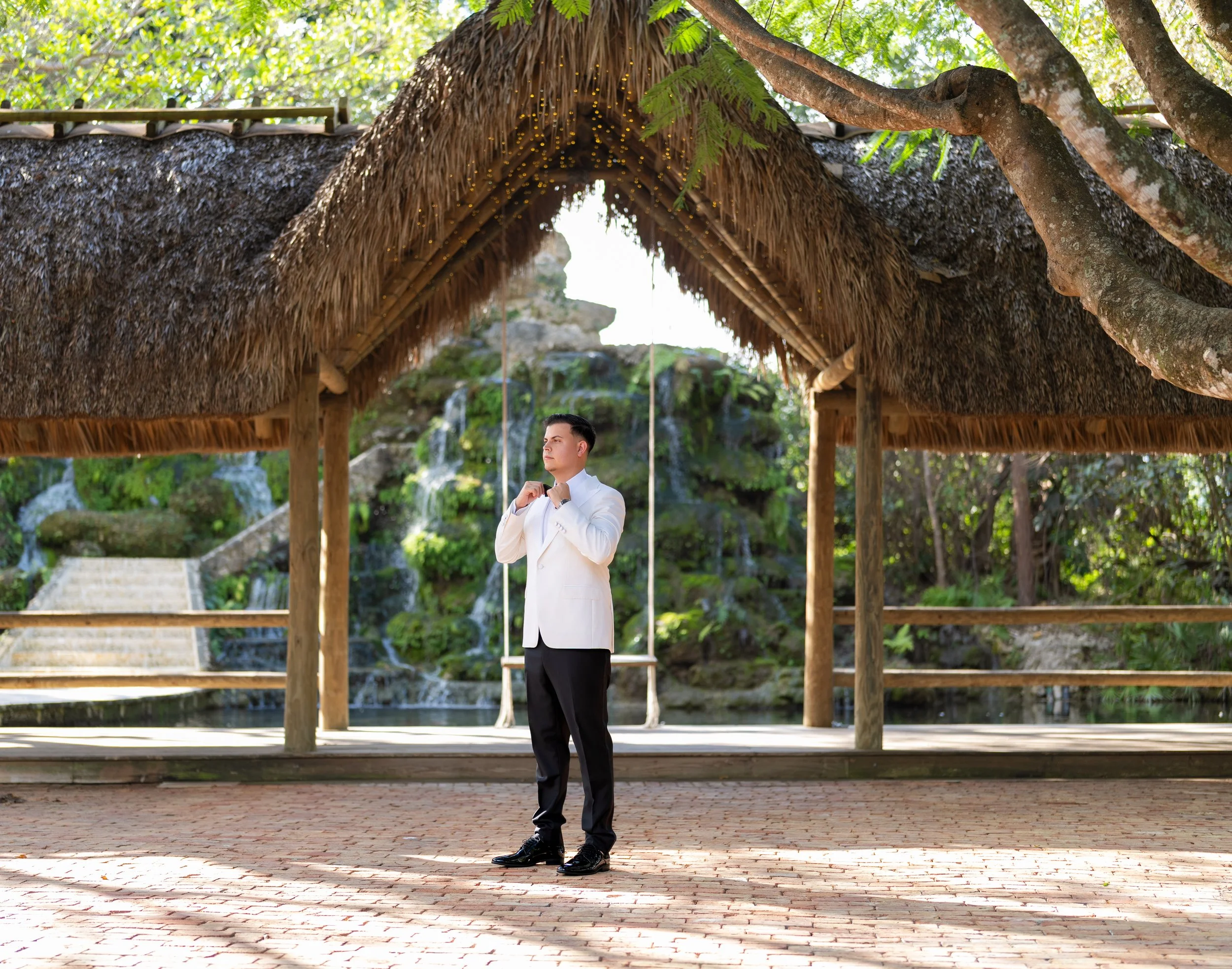 A man in a white tuxedo adjusting his bowtie under a thatched roof pavilion with a stone staircase and a waterfall in the background.