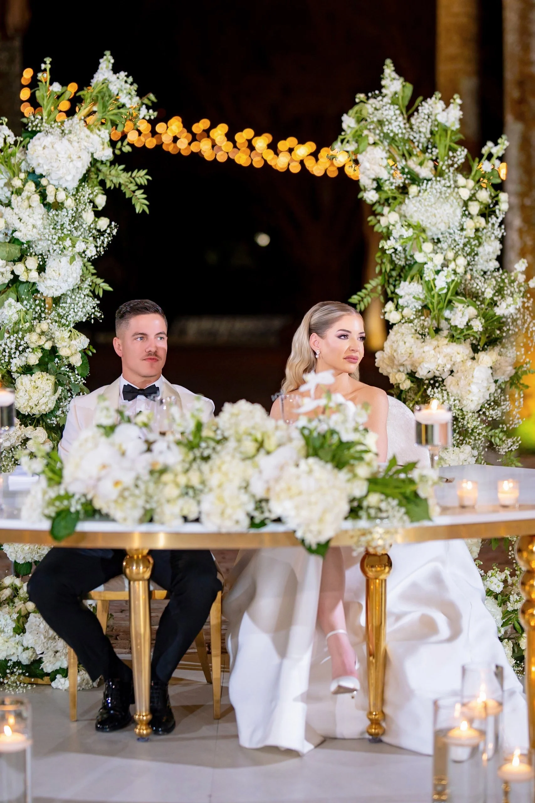 A wedding reception scene with a bride and groom sitting at a table adorned with white flowers and candles, with a floral arch behind them and string lights overhead.