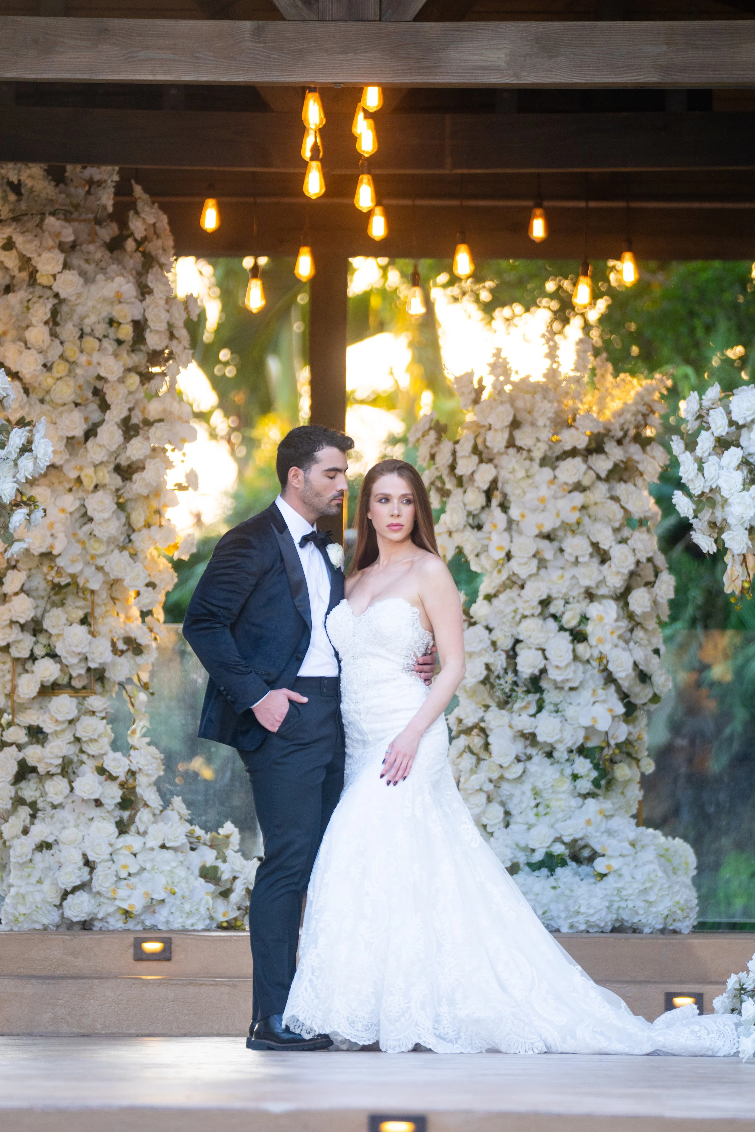 A bride and groom standing close together on a wedding stage decorated with white flowers and fairy lights at sunset.