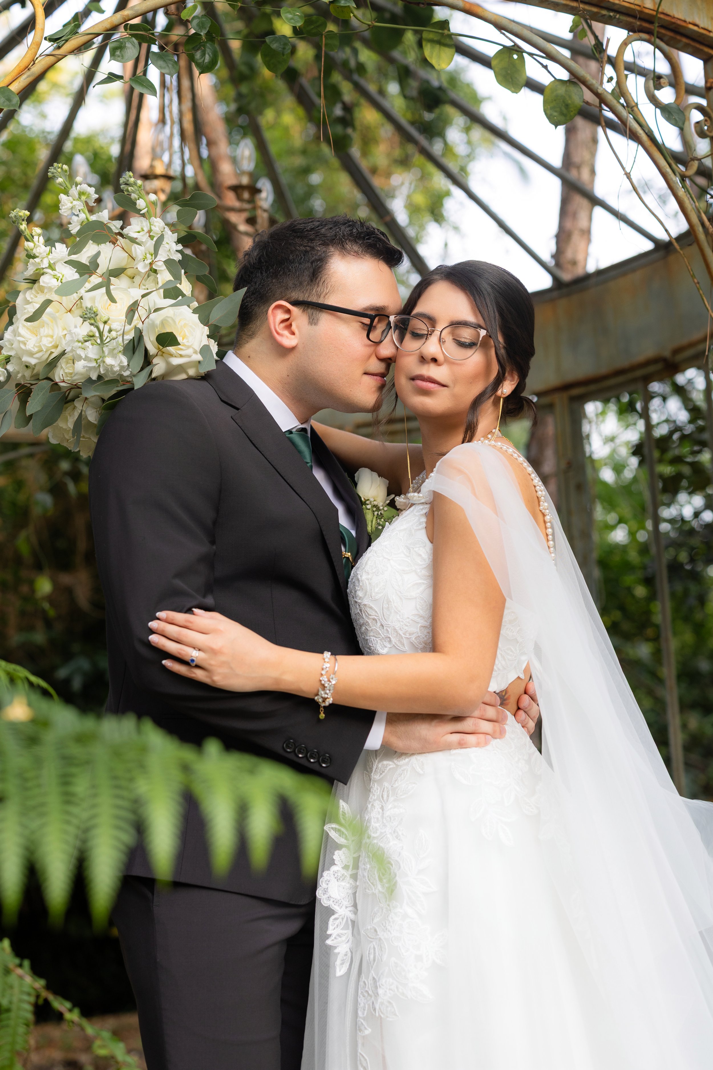 A bride and groom embrace each other outdoors during their wedding ceremony, surrounded by greenery and flowers.