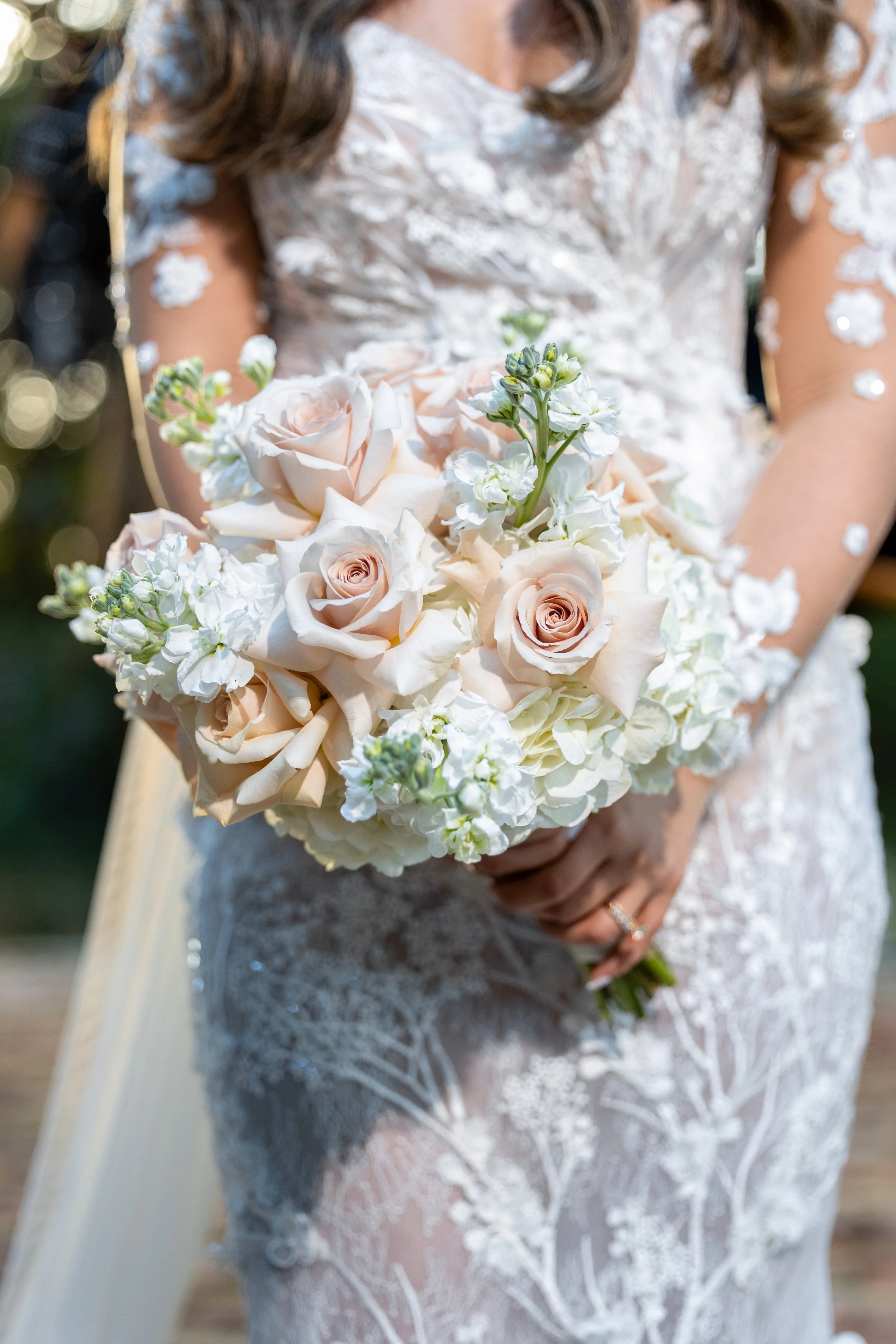 A bride in a white lace dress holding a bouquet of blush pink and white roses and other white flowers.