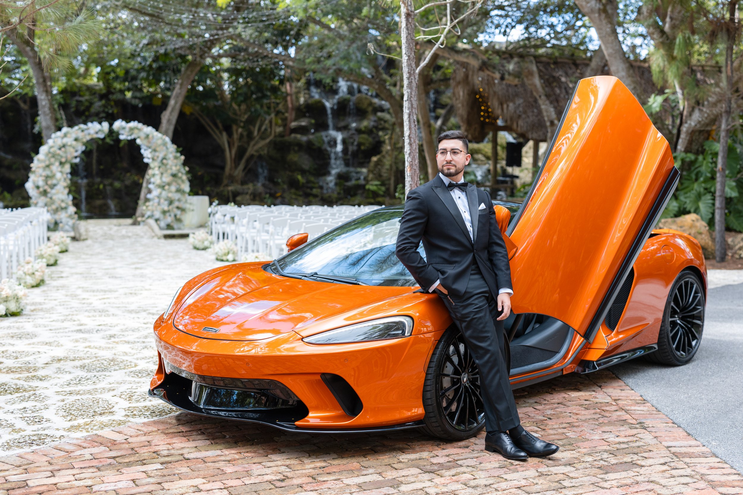 A man in a black tuxedo standing with one hand in his pocket leaning against an orange luxury sports car with gullwing doors partially open. He is at an outdoor wedding venue with white chairs, flowers, and a floral arch in the background.