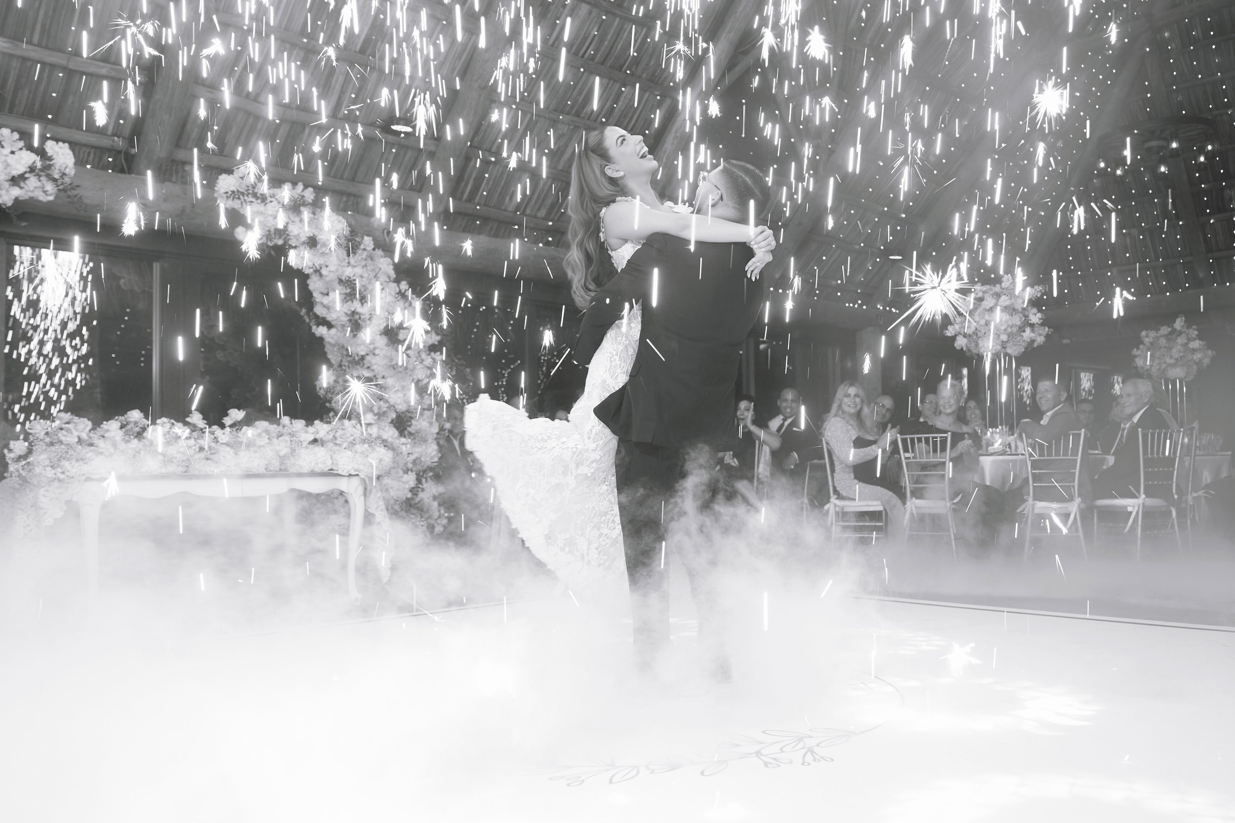 A woman in a wedding dress being lifted by a man at her wedding reception, with sparklers and fog creating a celebratory atmosphere.