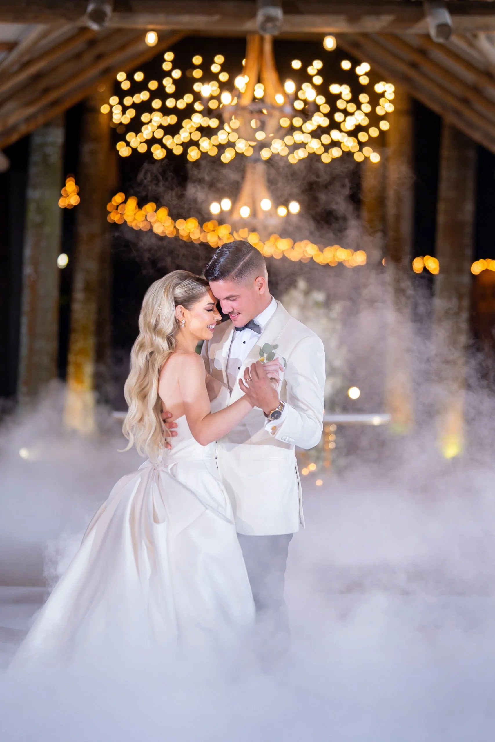 A bride and groom dance closely during their wedding reception, with warm lighting and chandelier in the background, surrounded by fog.
