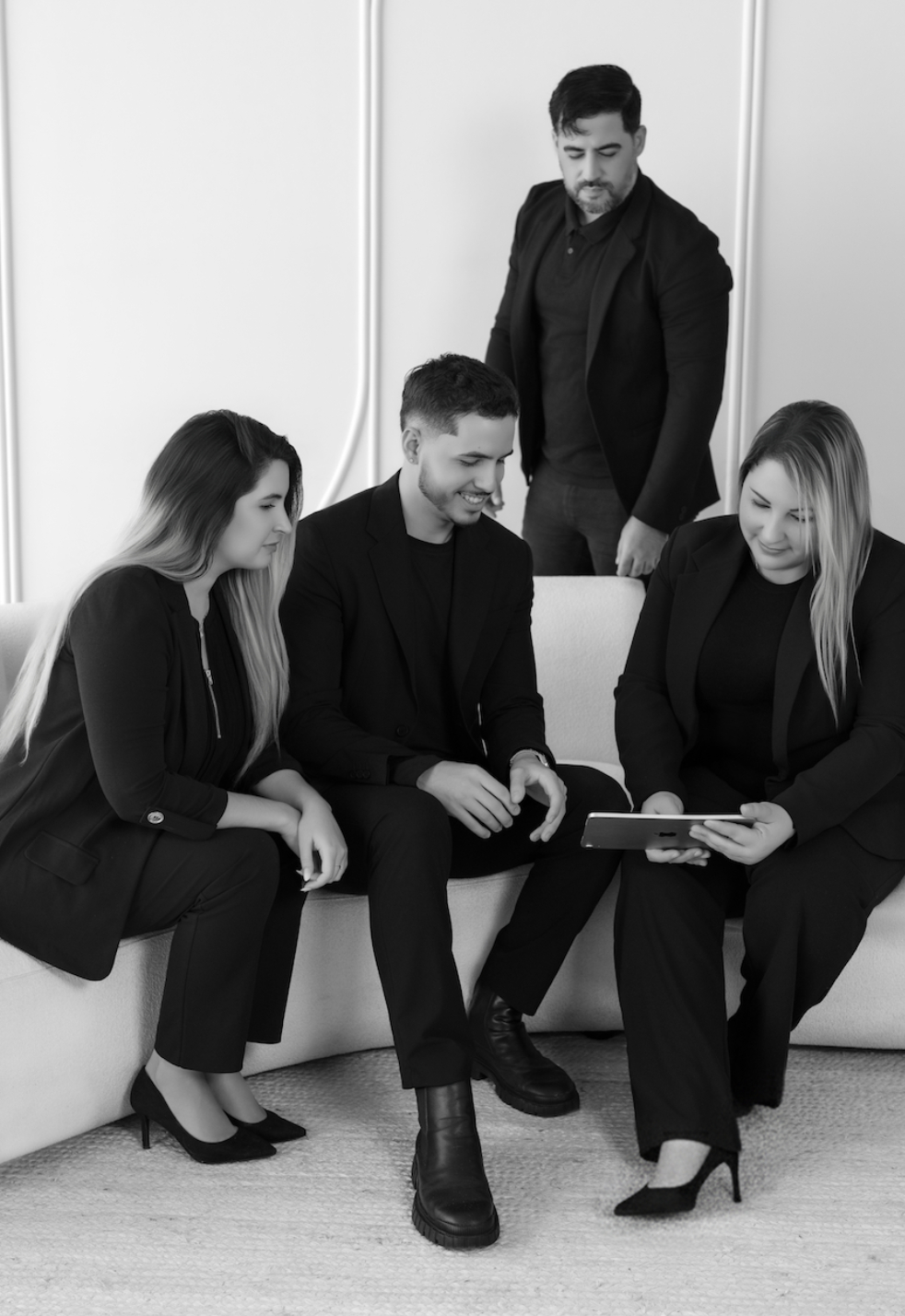 Four young professionals in formal business attire sitting and standing around a tablet, engaging in a discussion in a modern office setting.
