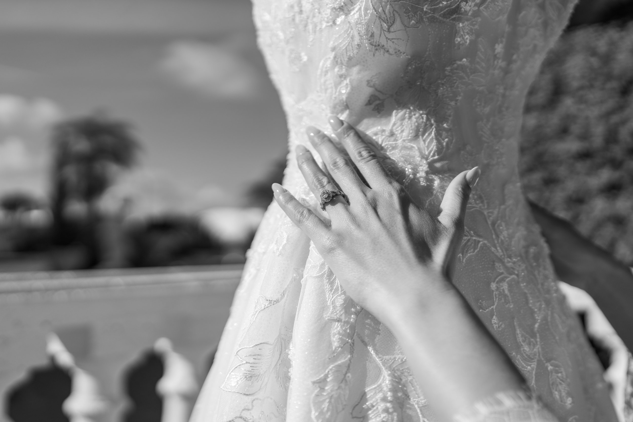 A close-up of a woman's hand with a ring, resting on her wedding dress outdoors on a sunny day. The background shows trees and a fence, slightly out of focus.