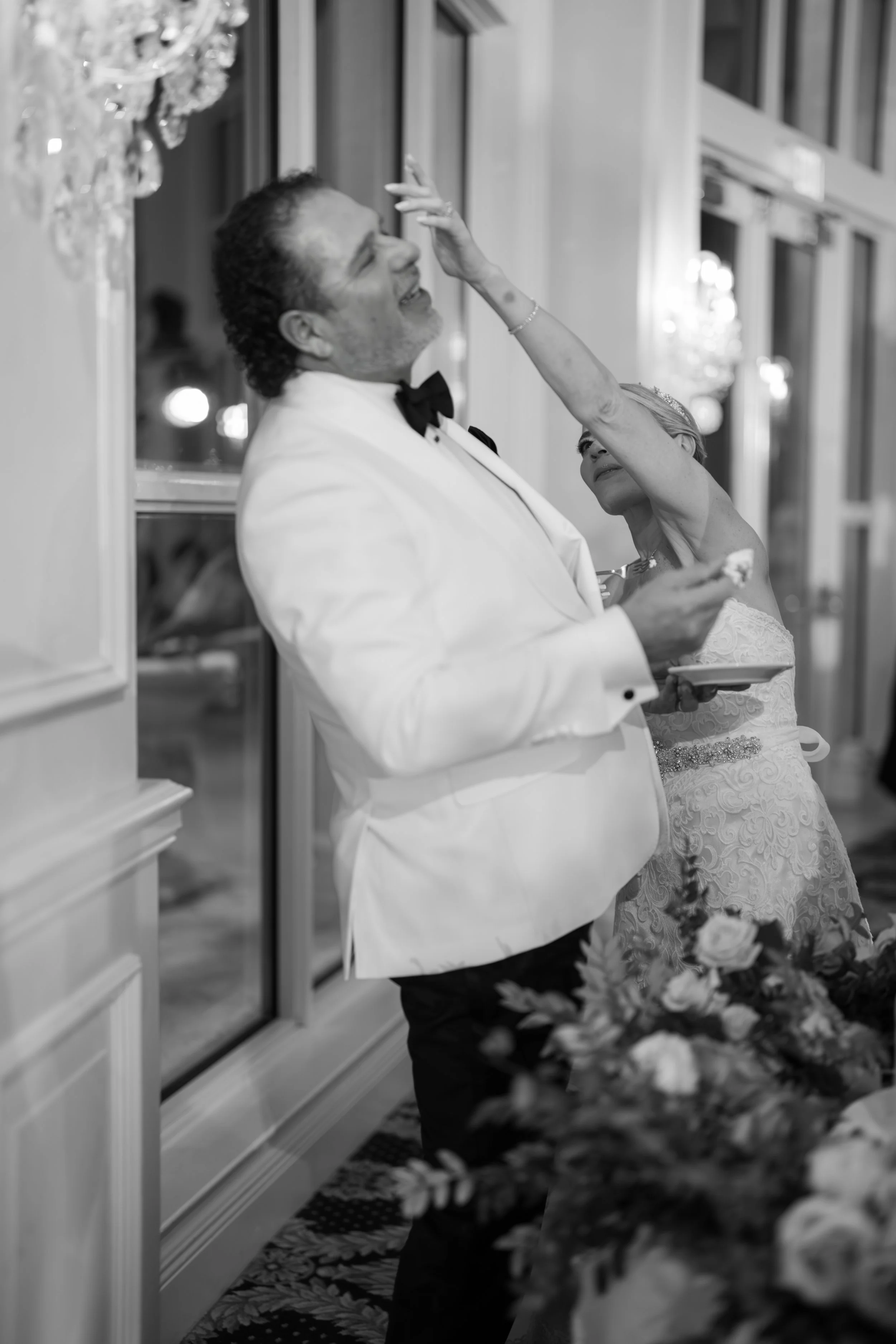 A groom and bride sharing a moment at their wedding reception, with the groom wearing a tuxedo and the bride in a wedding dress, inside a decorated banquet hall.
