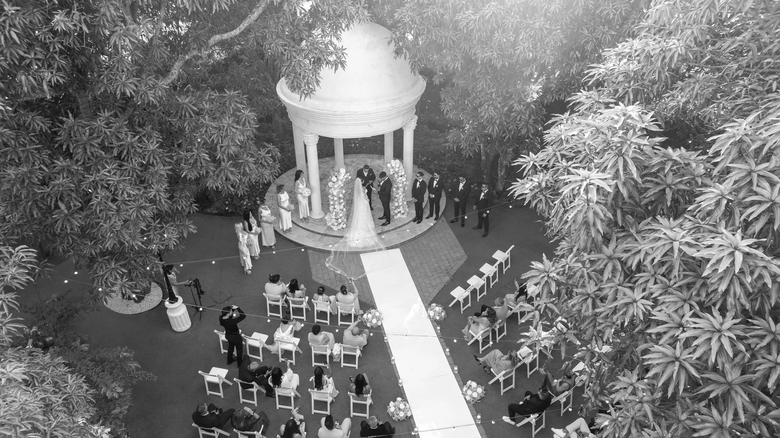 A black and white aerial view of an outdoor wedding ceremony in a garden, with guests seated on white chairs facing a small circular altar decorated with flowers, beneath a canopy supported by columns, surrounded by tall trees.