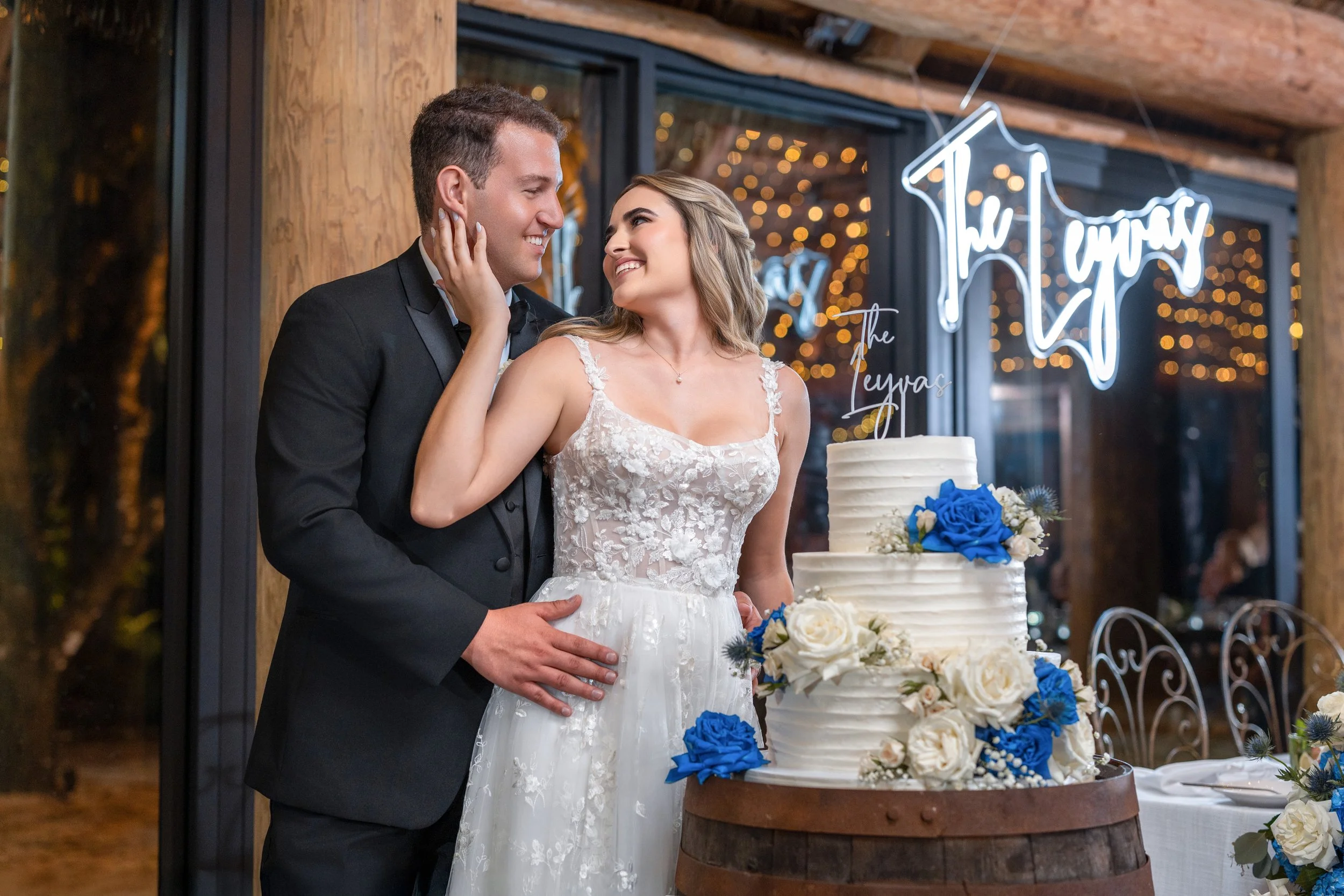 A bride and groom share a moment by a wedding cake decorated with blue and white flowers, inside a decorated venue with a neon sign reading 'This Happy' in the background.
