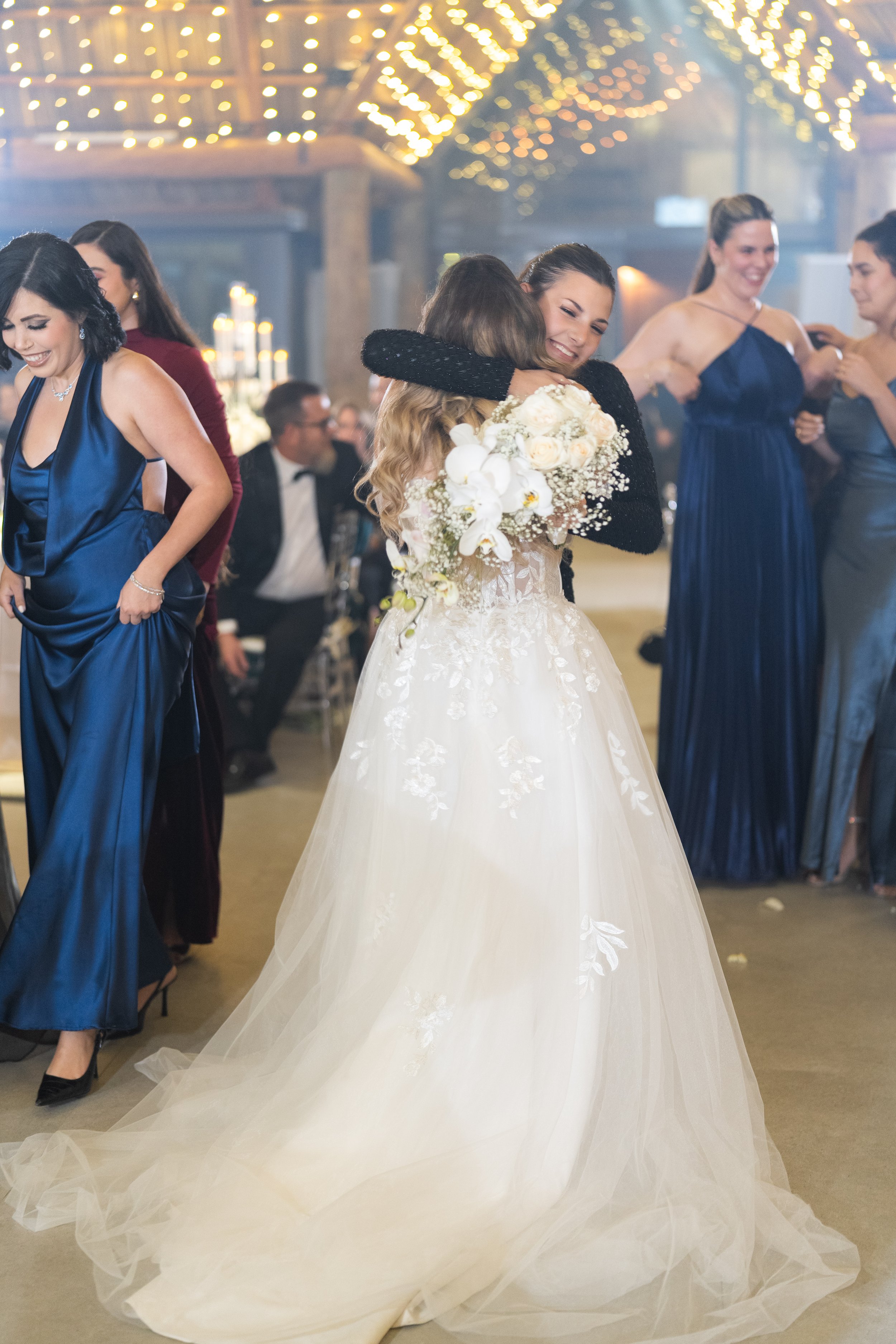 Bride in a white wedding gown hugging a woman in a black dress at a wedding reception with guests in formal attire, warm lighting, and fairy lights overhead.