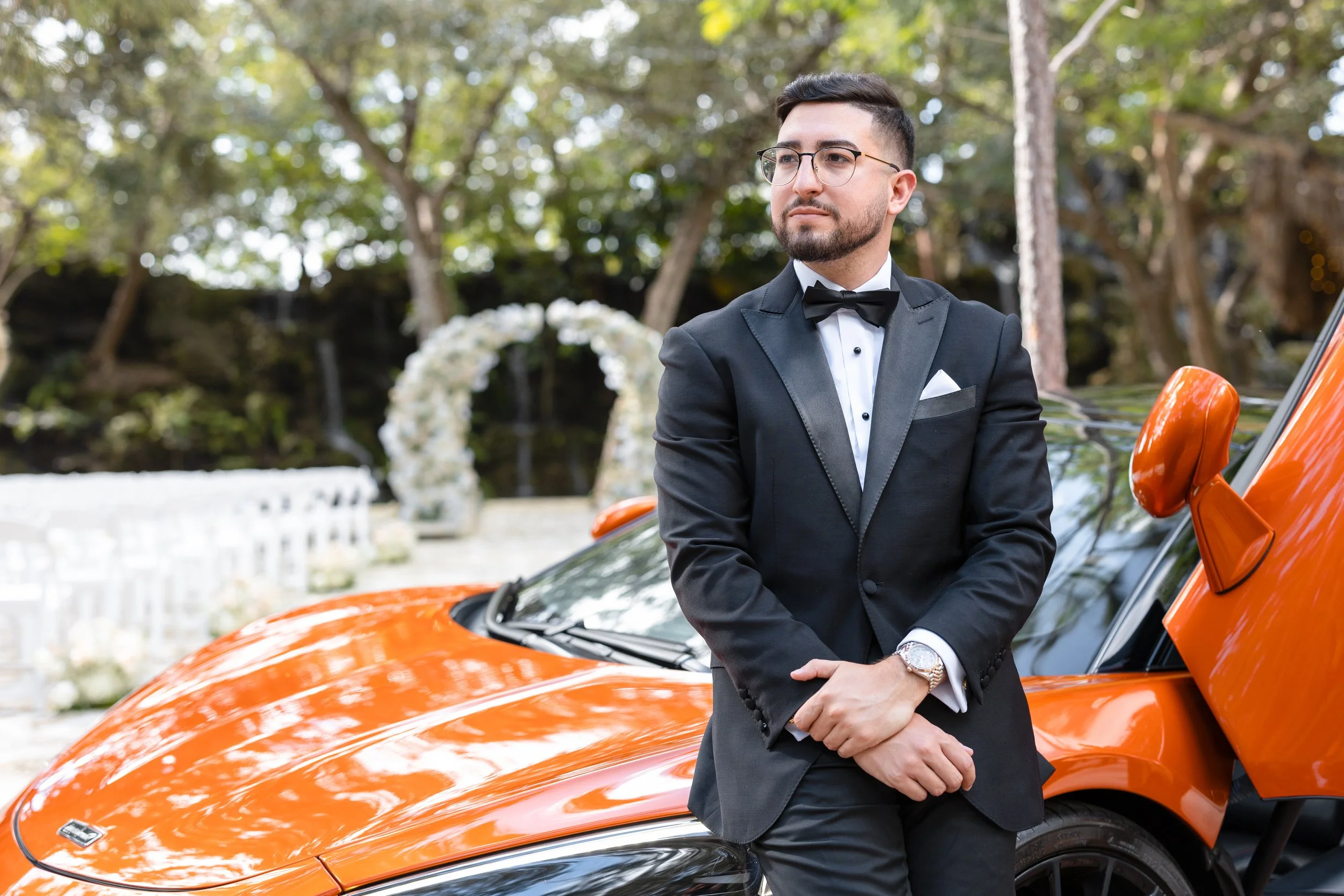A man in a tuxedo standing next to an orange sports car at an outdoor wedding ceremony with a floral arch in the background.
