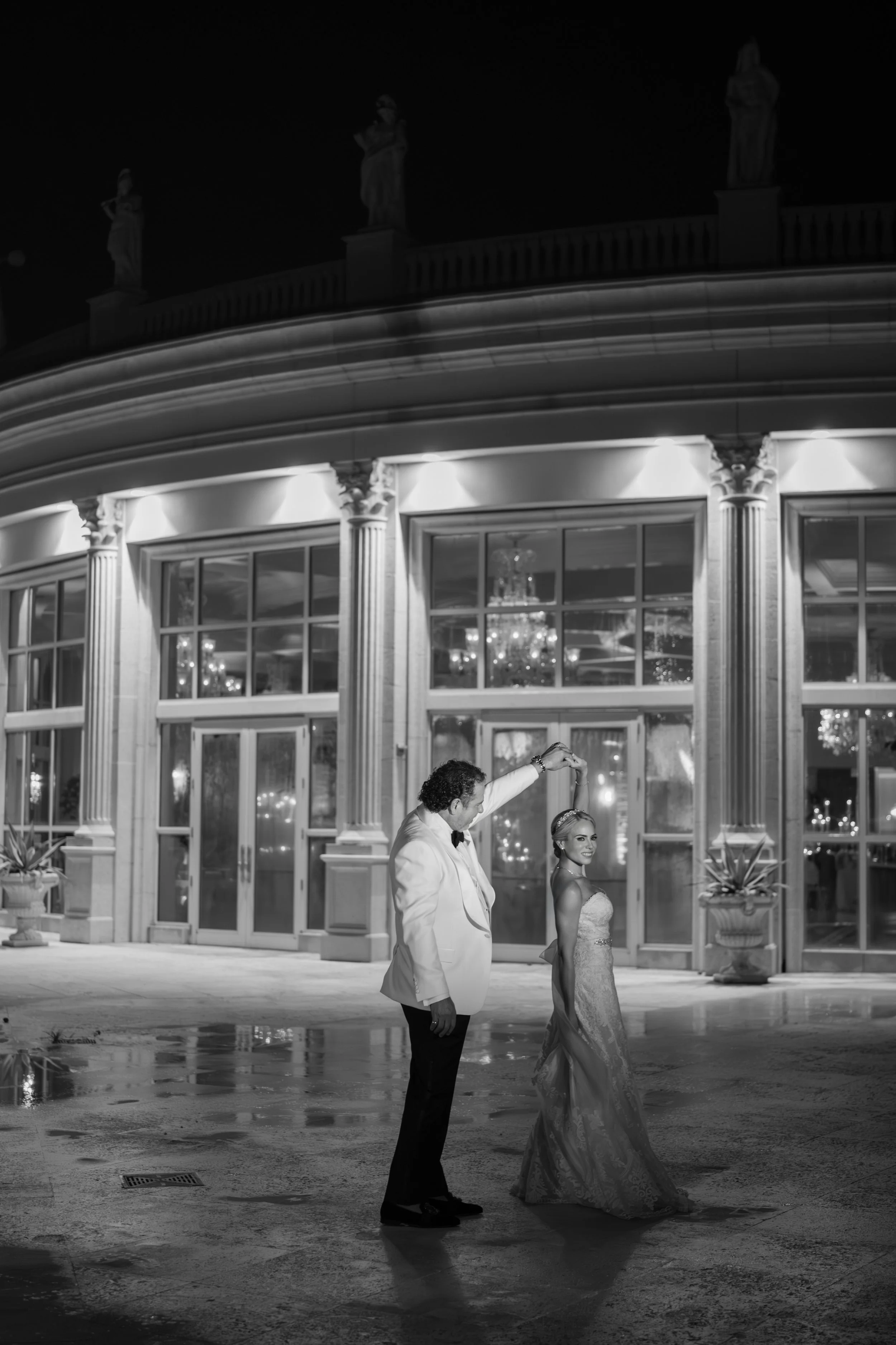 A black and white photo of a bride and groom dancing outdoors at night in front of a grand building with tall windows and decorative columns. The groom is holding the bride's hand as she spins.