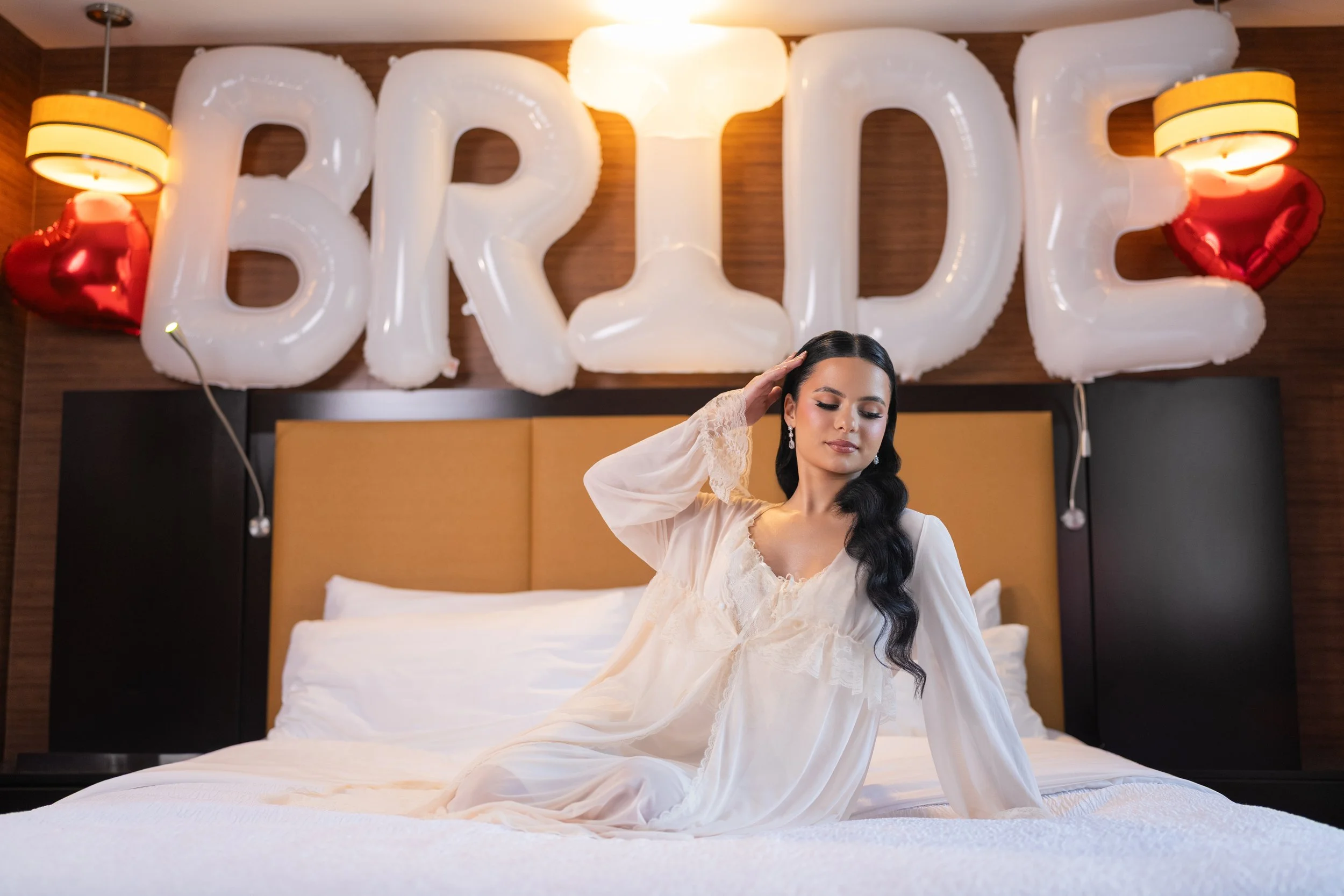 A woman in a white dress sitting on a bed with large 'BRIDE' balloon letters in the background, decorated with heart-shaped balloons and warm lighting.