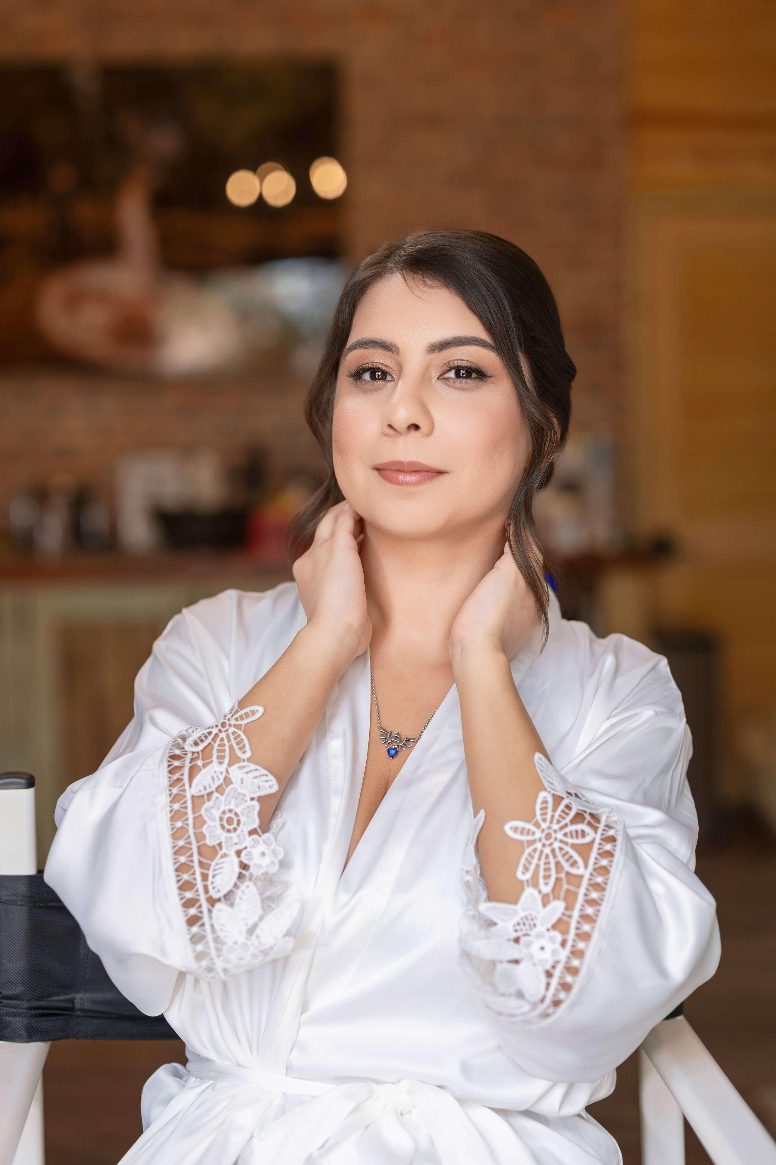 A young woman with dark hair styled in loose waves, wearing a white satin robe with lace embroidered sleeves, sitting in a cozy indoor space with warm brick walls and blurred background decor.