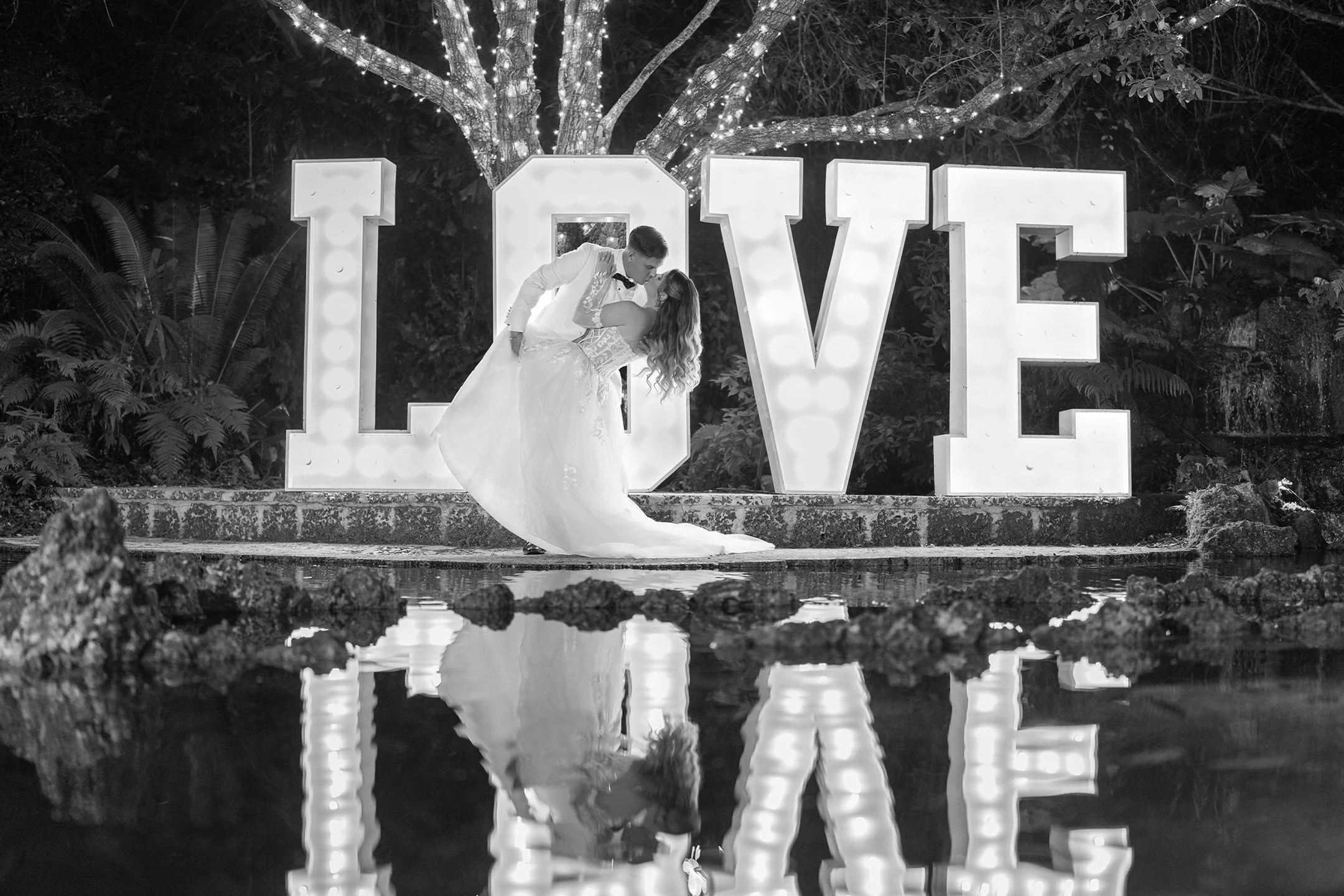 A bride and groom sharing a dance in front of large illuminated letters spelling LOVE, with a tree decorated with lights in the background, reflected in a pond in the foreground.