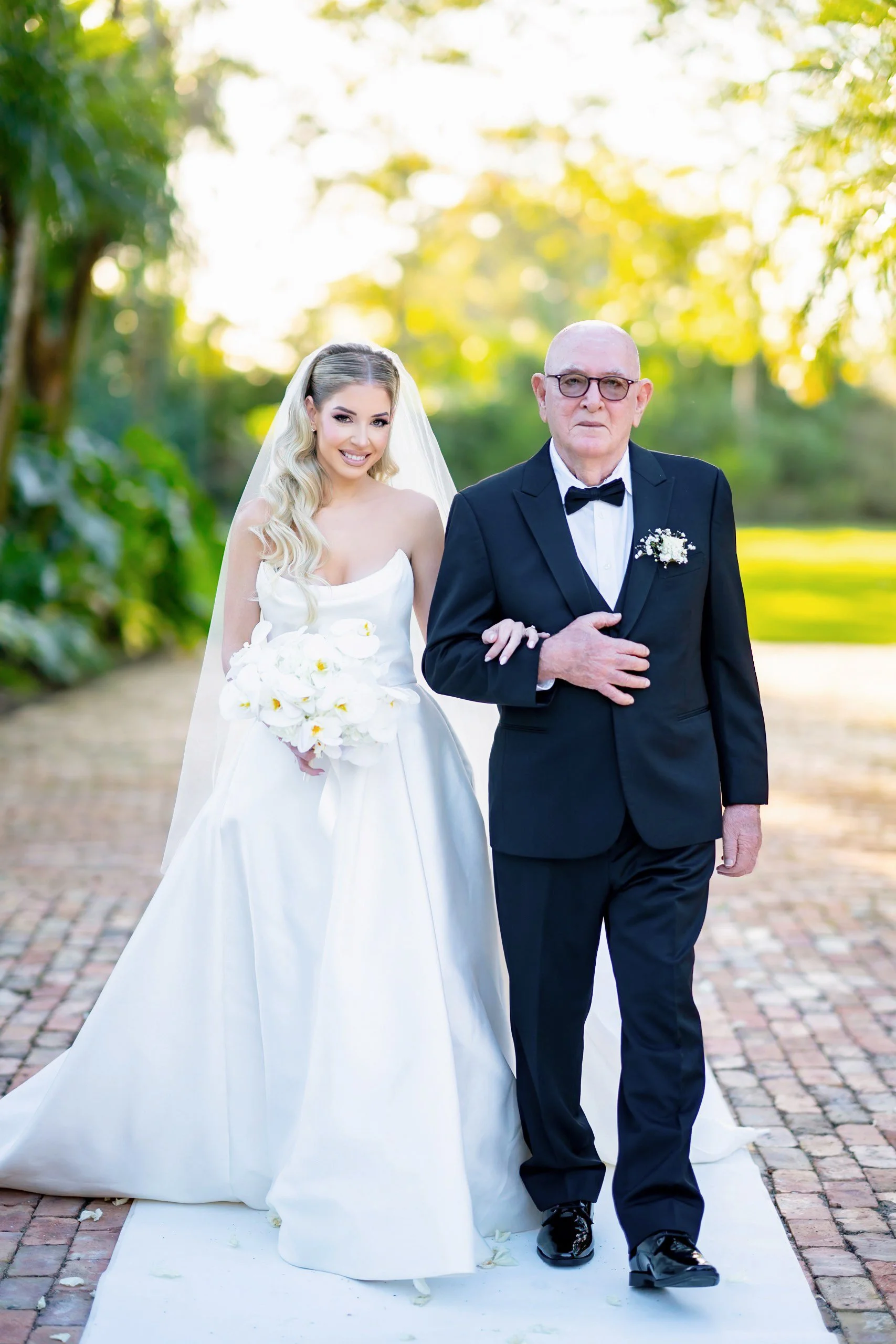 A bride in a white wedding dress and veil holding a bouquet of white calla lilies walking down the aisle with an older man in a black tuxedo, during an outdoor wedding ceremony.