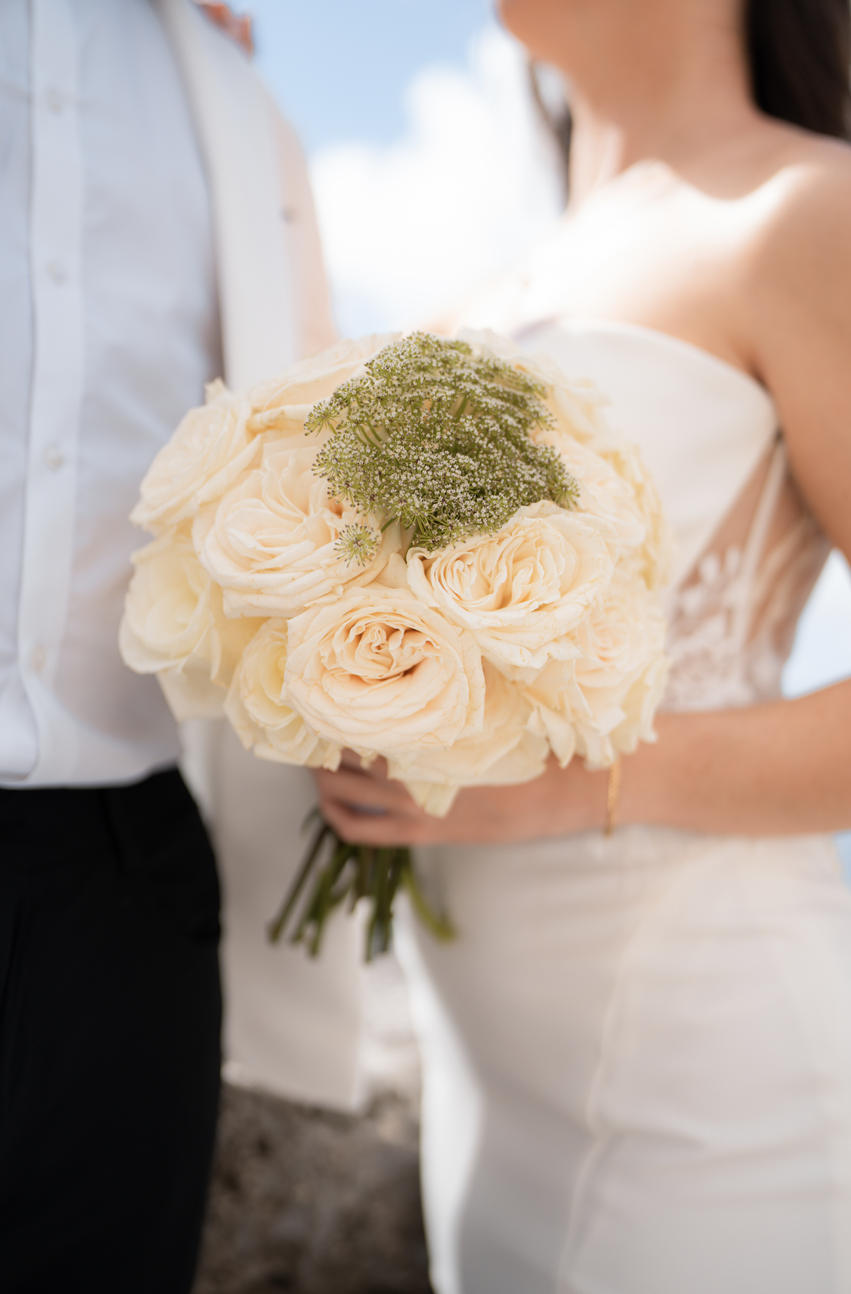 A wedding bride holding a bouquet of cream-colored roses with greenery, standing with a groom in formal attire outdoors on a sunny day.