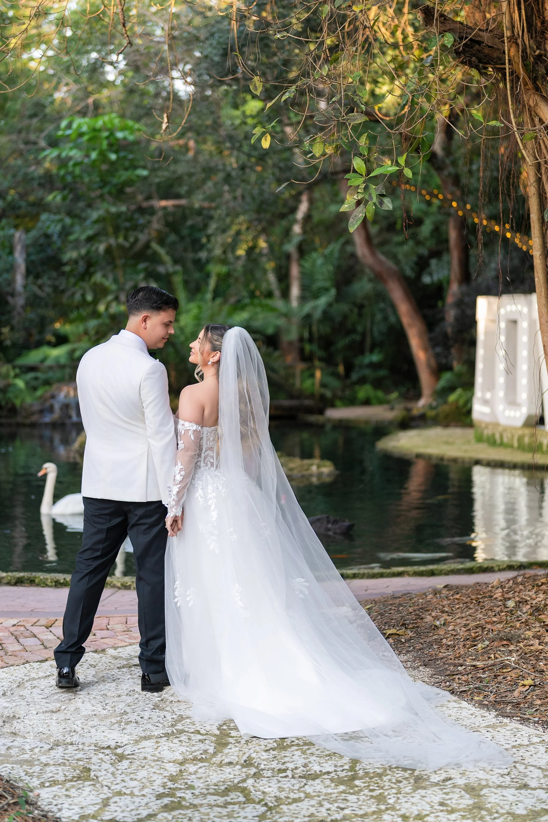 A bride and groom holding hands and looking at each other outdoors, in a natural setting with a pond and swan in the background, during their wedding ceremony.