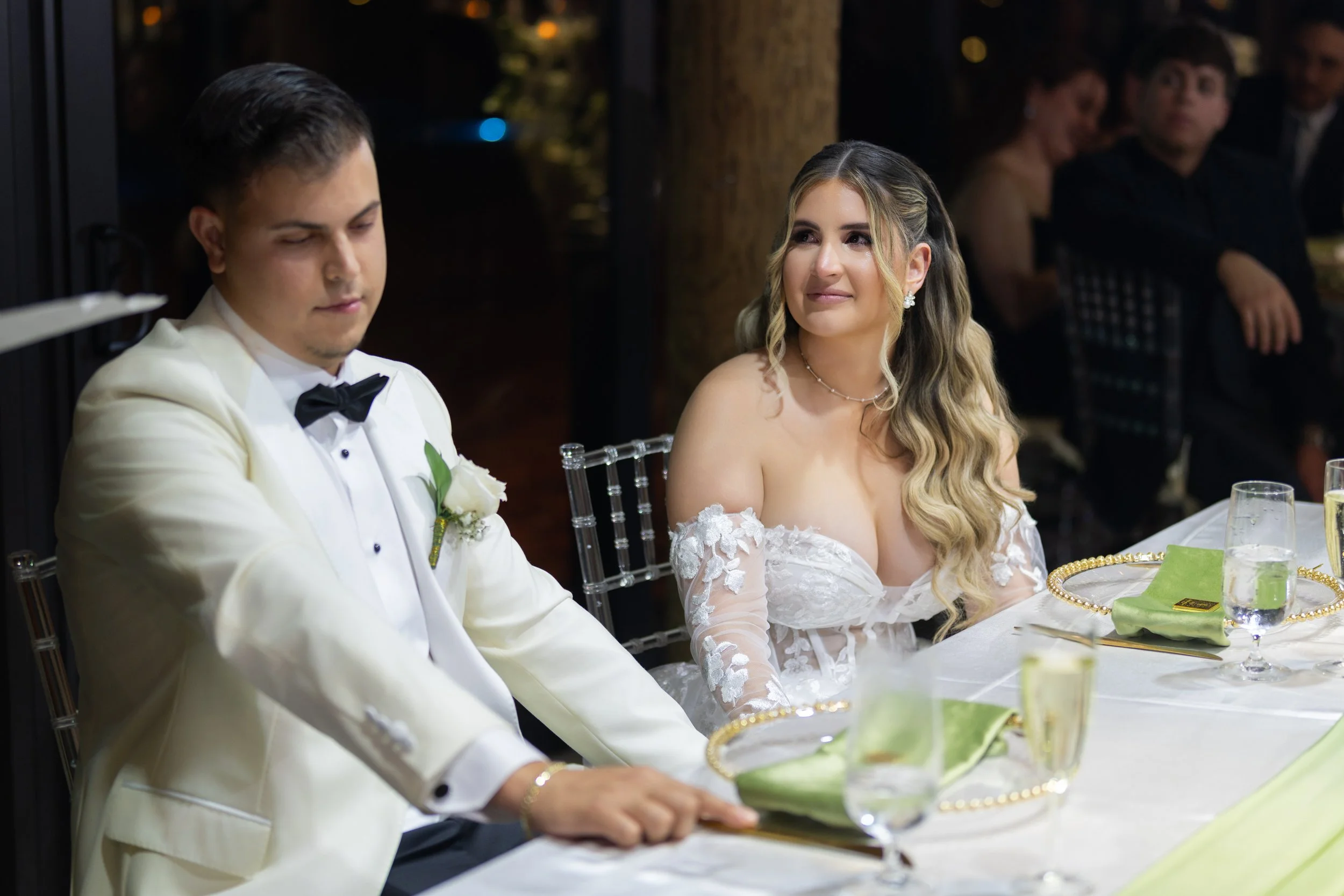 A bride and groom sitting at a wedding reception table, with guests in the background. The bride has long wavy hair and is wearing an off-the-shoulder white wedding gown. The groom is wearing a white tuxedo with a black bow tie.