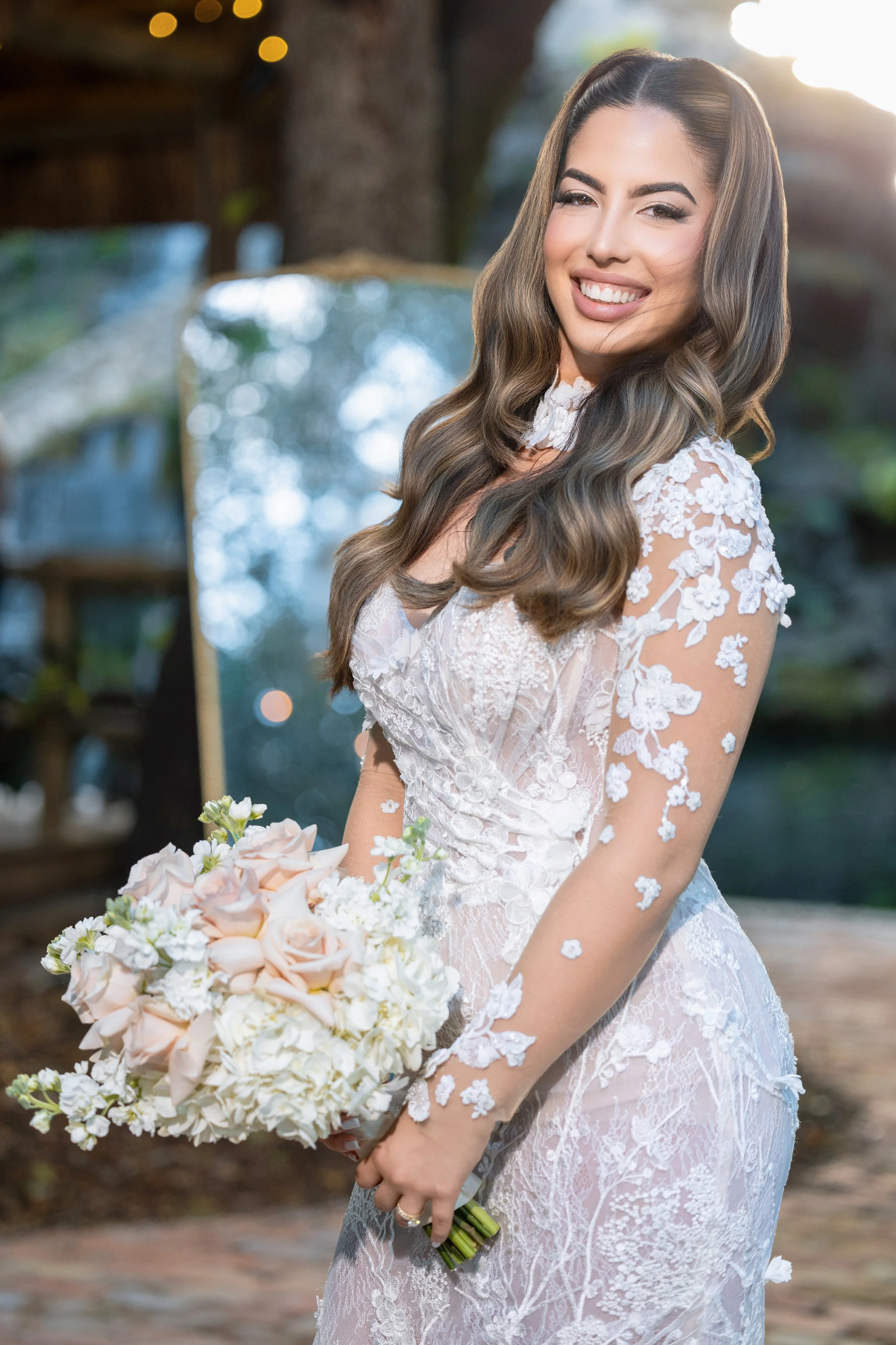 A smiling woman in a lace wedding dress holding a bouquet of pink and white flowers outdoors.