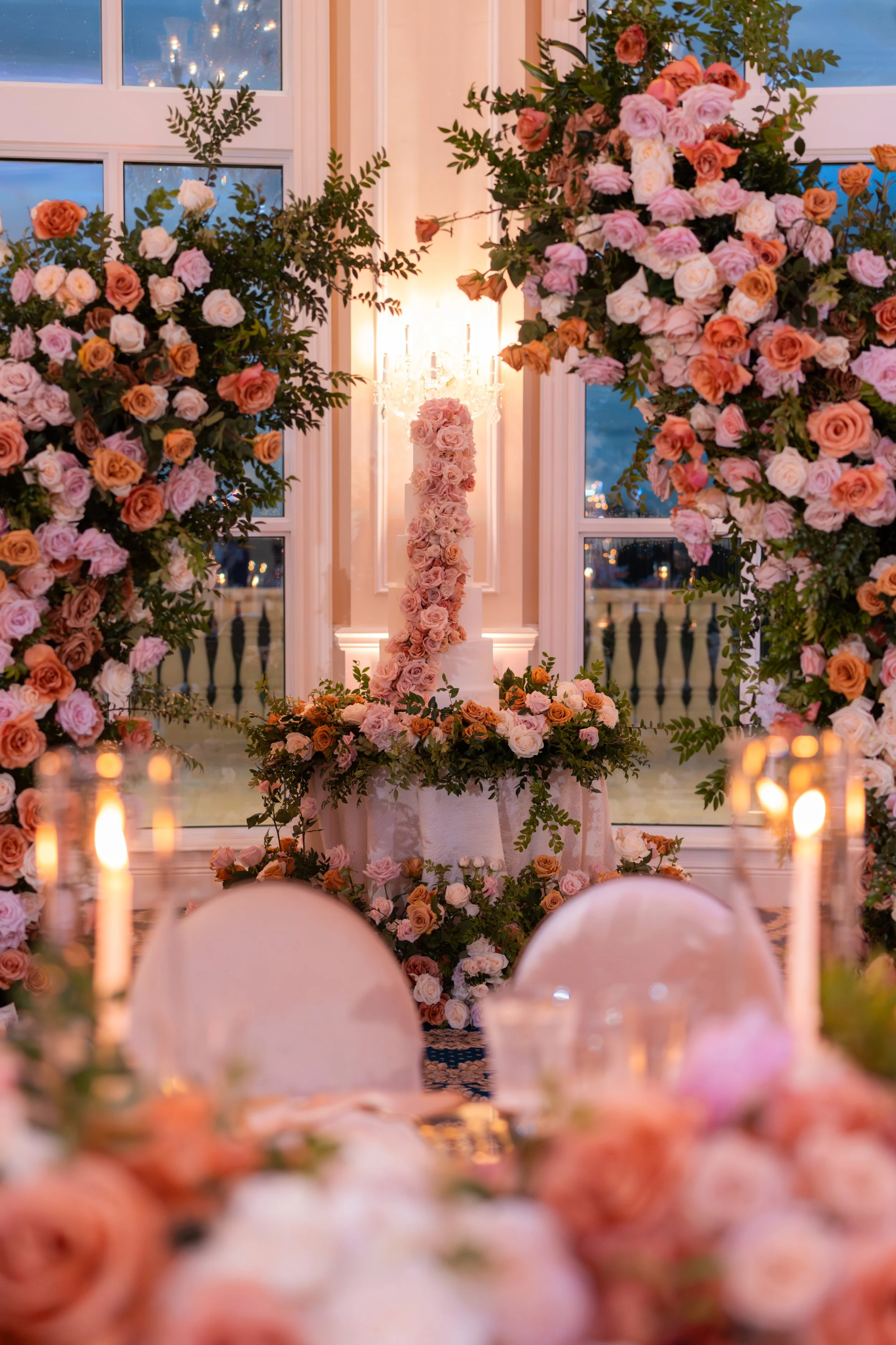 Elegant wedding decor with pink, peach, and white roses, floral arches, and a tiered cake adorned with flowers, set by large windows and lit by candlelight.