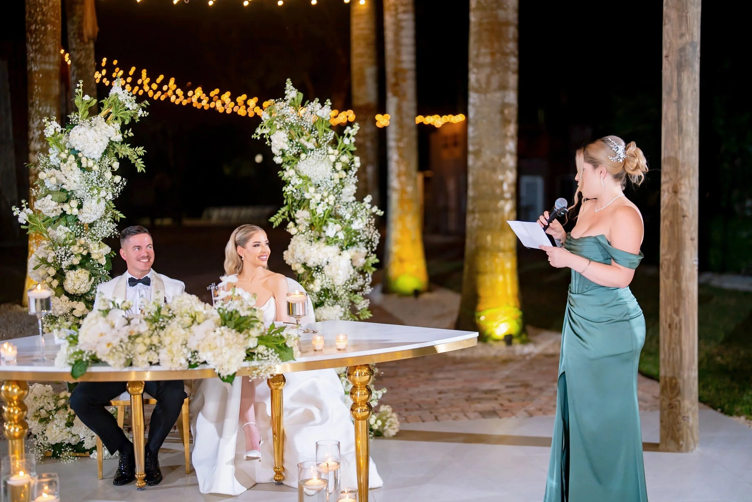 A bride and groom sitting at a wedding reception table with floral decorations, while a woman gives a speech or toast holding a microphone and reading from a paper, outdoors at night with string lights and lit candles.
