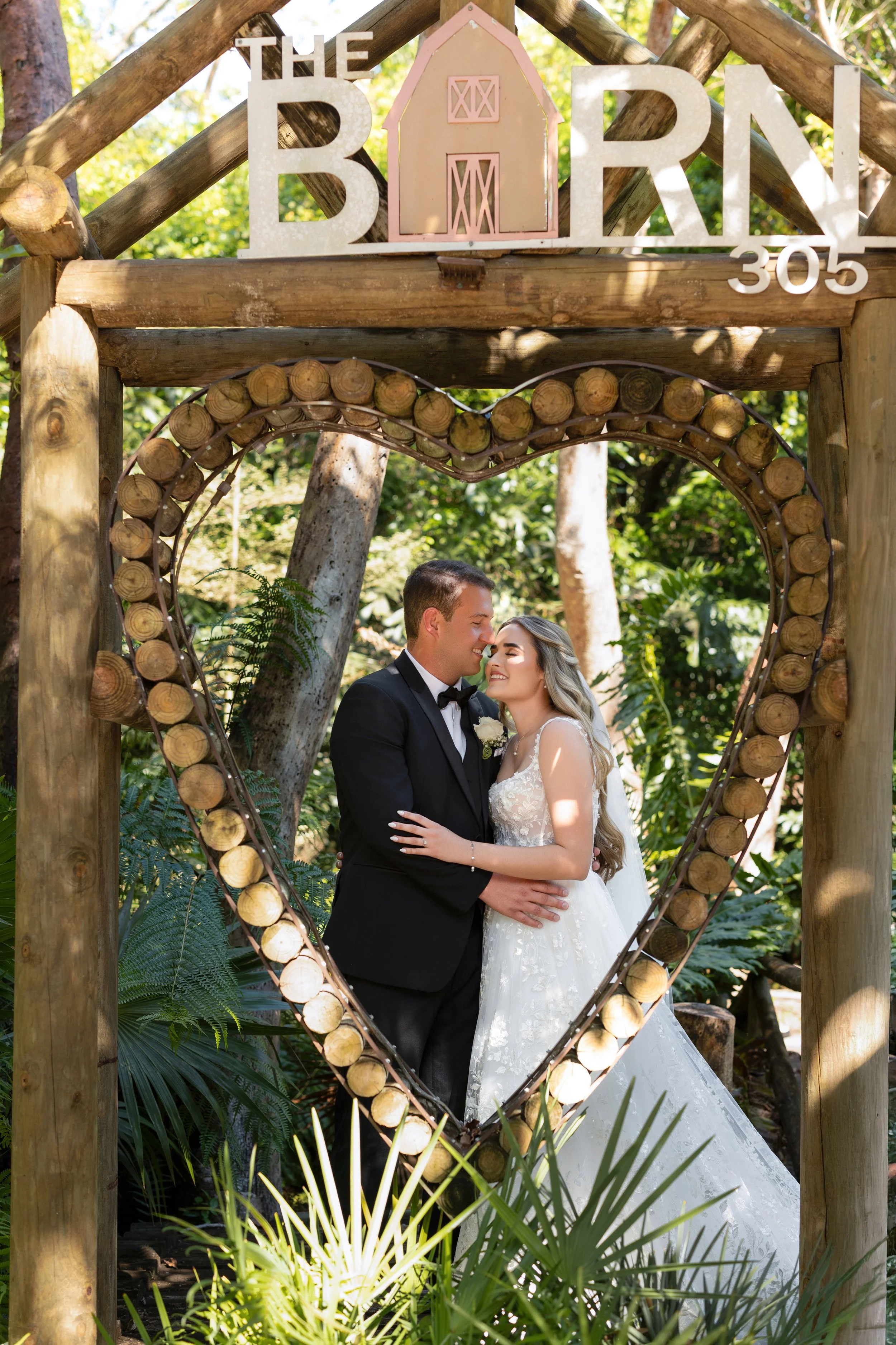 A bride and groom smiling and embracing inside a wooden heart-shaped frame outdoors surrounded by green plants. The scene is decorated with a sign that says 'THE BARN 305' and a pink barn-shaped decoration.