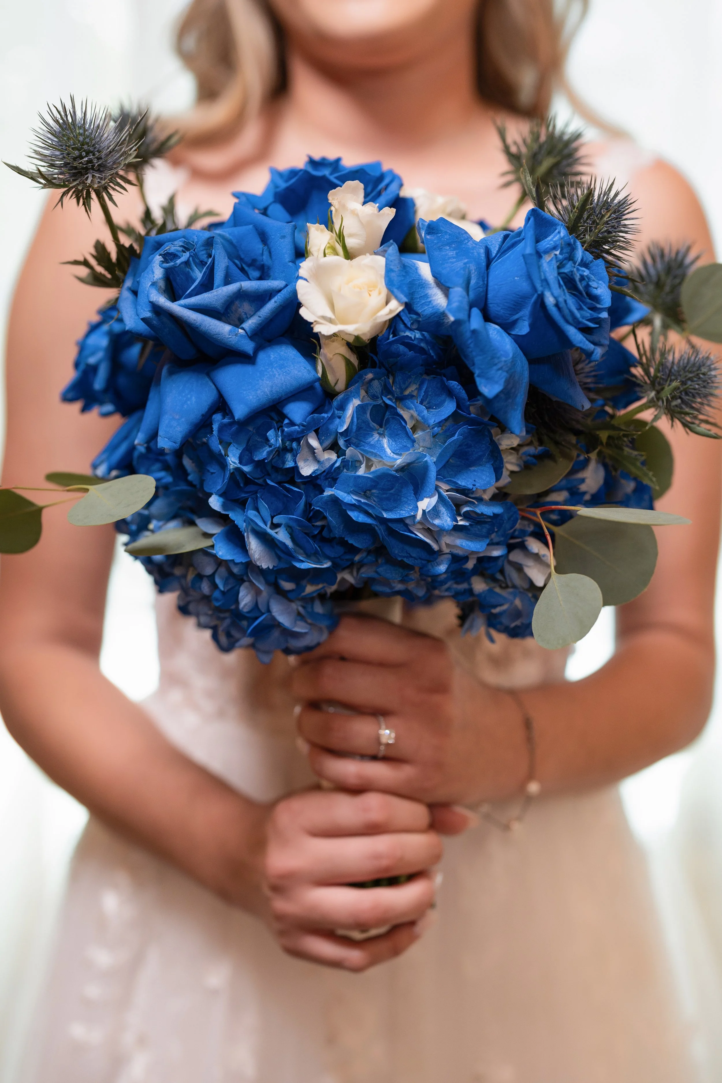 A bride holding a bouquet of blue hydrangeas, white roses, eucalyptus leaves, and spiky purple thistles, wearing a wedding ring and bracelet.