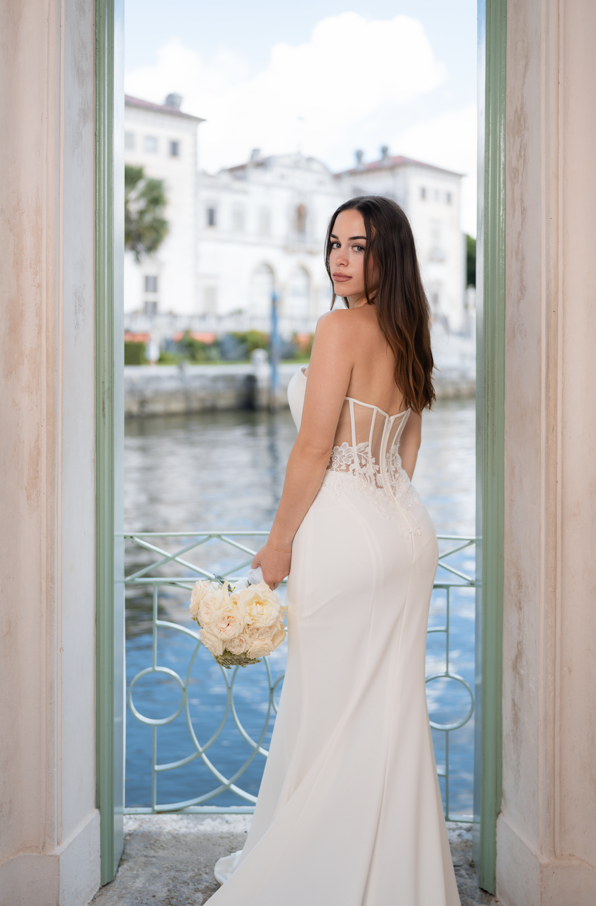 A bride in a white wedding gown holding a bouquet, standing in a doorway by a canal with white buildings and a blue sky in the background.