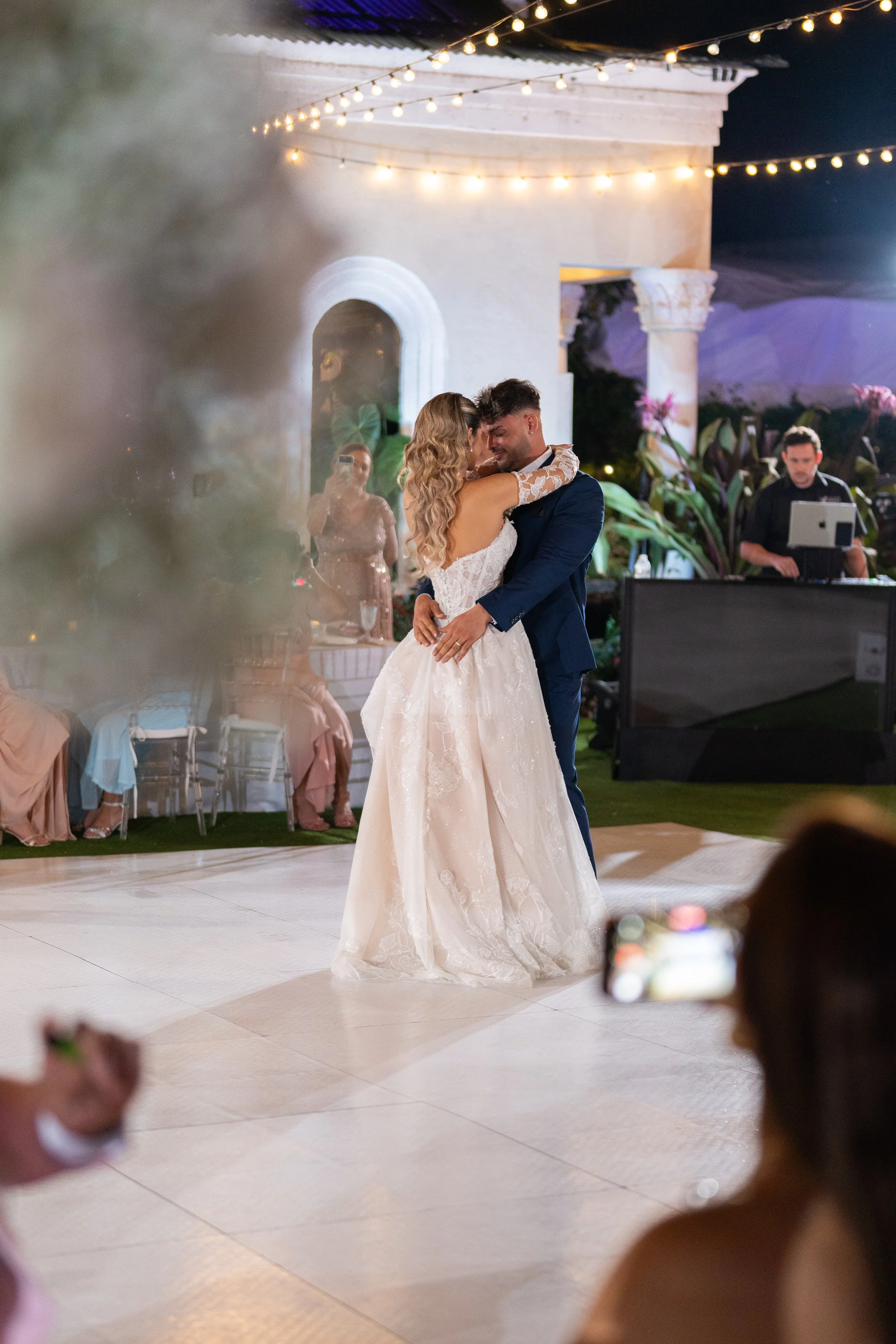 Bride and groom dancing at their wedding reception under string lights, with guests watching and a DJ in the background.