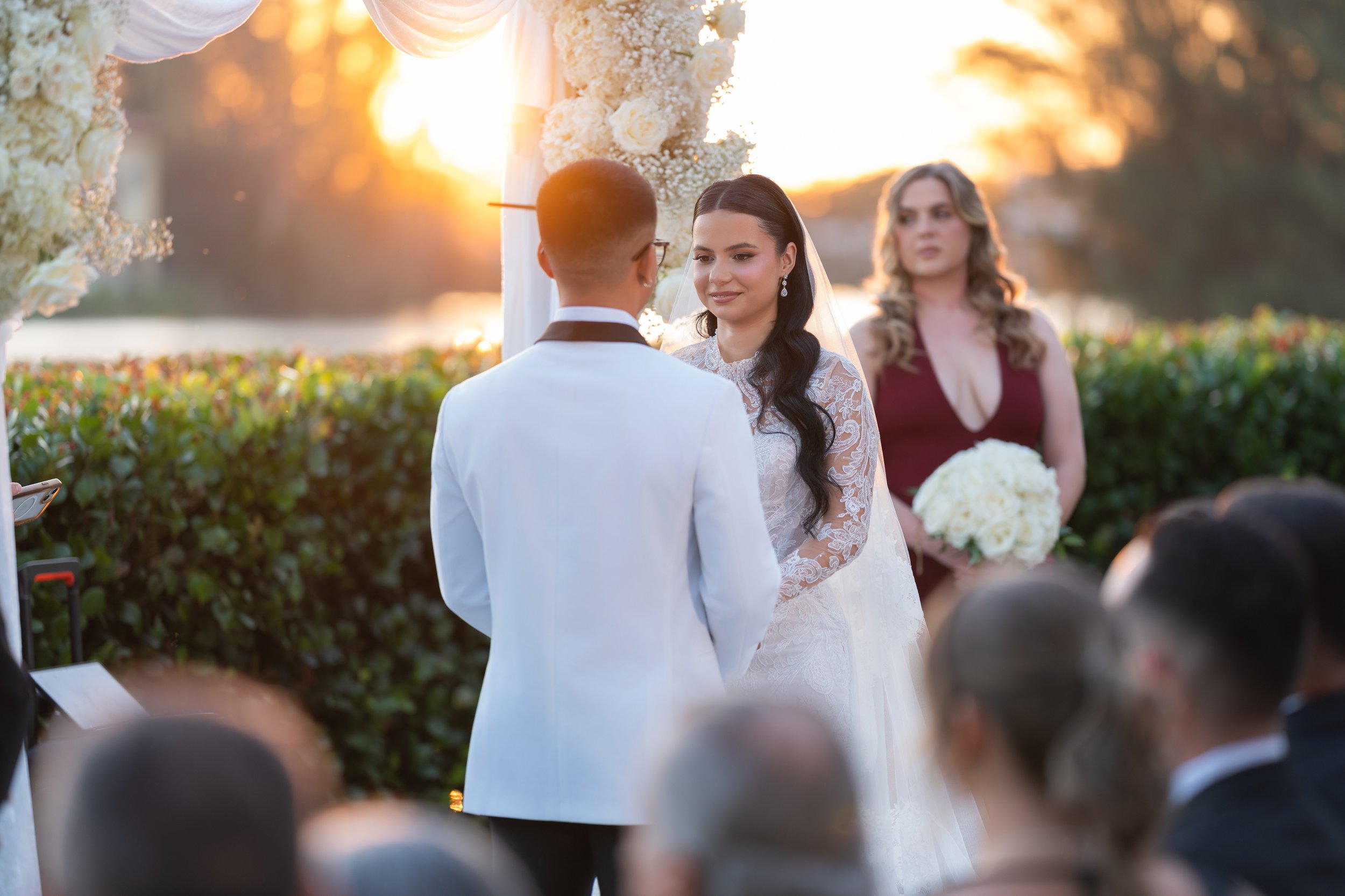 A wedding ceremony outdoors during sunset with a bride and groom facing each other, a woman holding a bouquet of white flowers standing behind them, and guests seated in front.