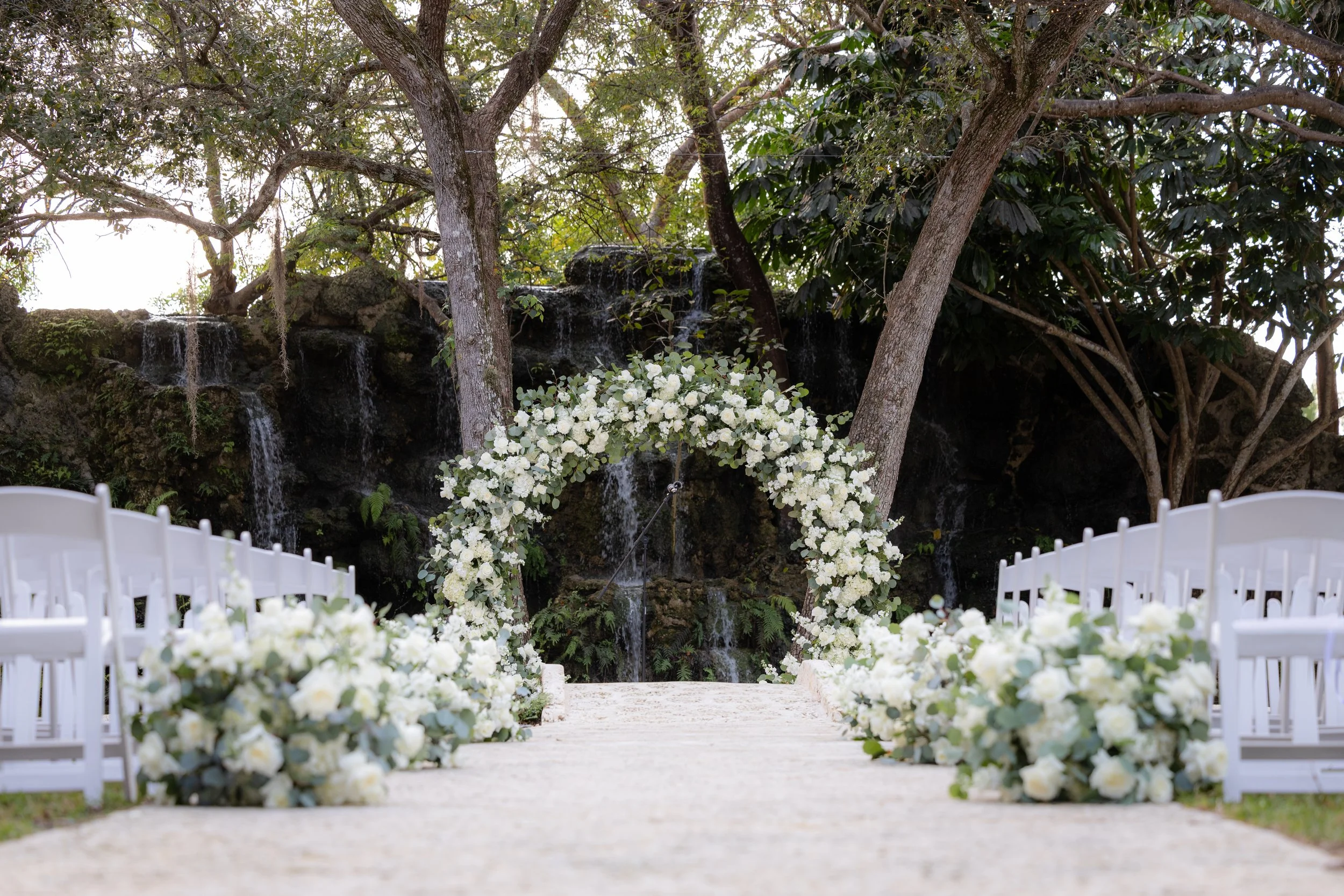 Outdoor wedding setup with white floral arch, surrounded by white chairs, against a backdrop of trees and waterfalls.