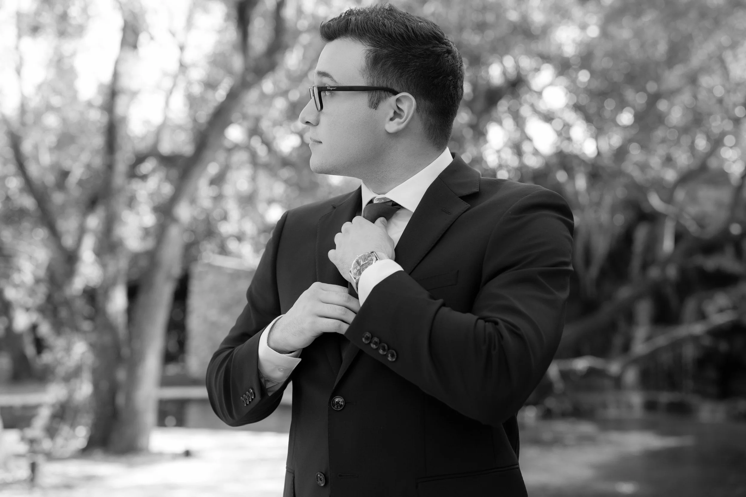 A man in a black suit and glasses adjusting his tie outdoors, with trees in the background.