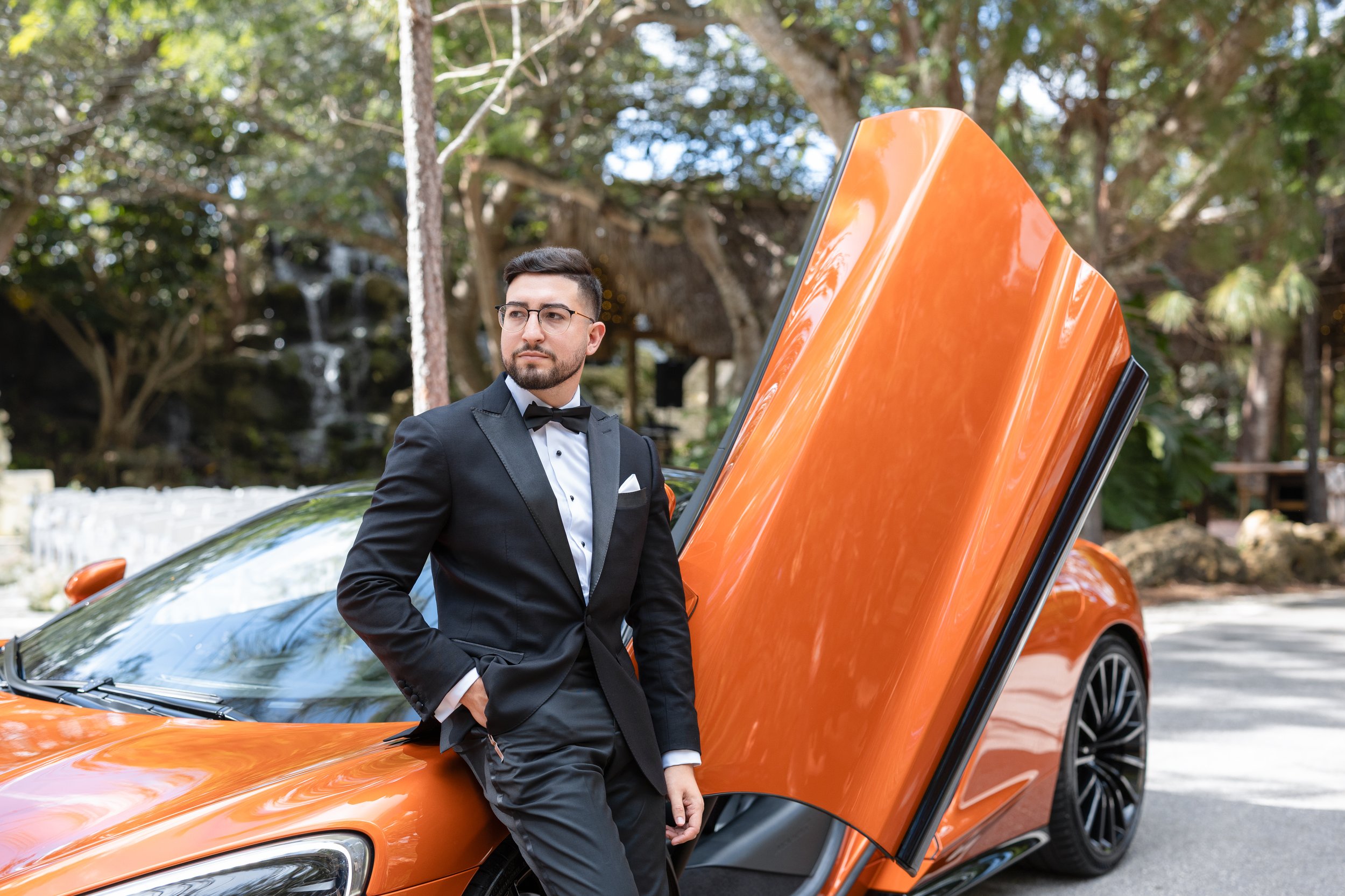 A man in a tuxedo leaning against an orange luxury sports car with scissor doors, outdoors with trees and a waterfall in the background.