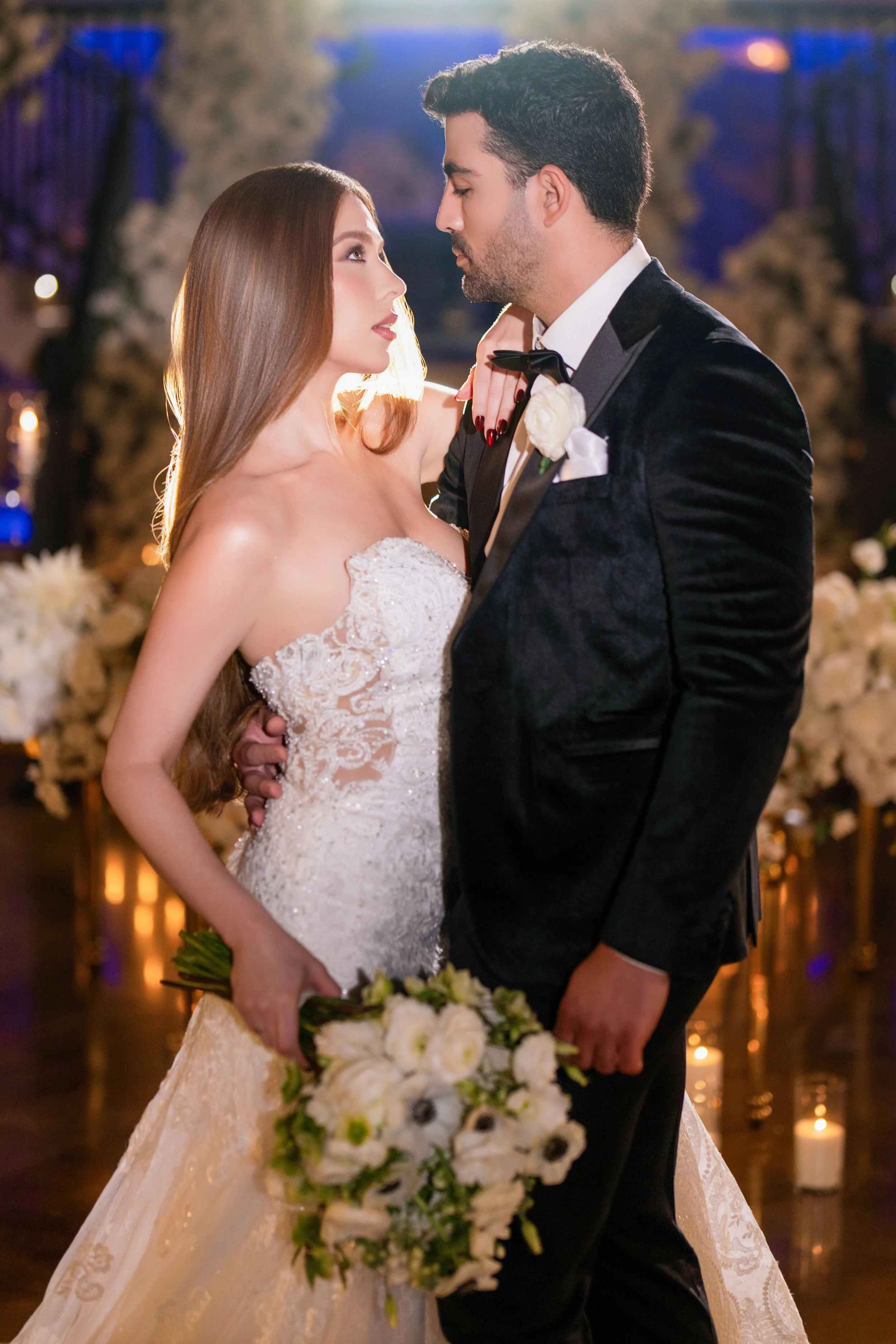 A bride and groom sharing a romantic moment during their wedding reception, with the bride holding a bouquet of white flowers and the groom wearing a black tuxedo with a white boutonniere, surrounded by floral decorations and candles.